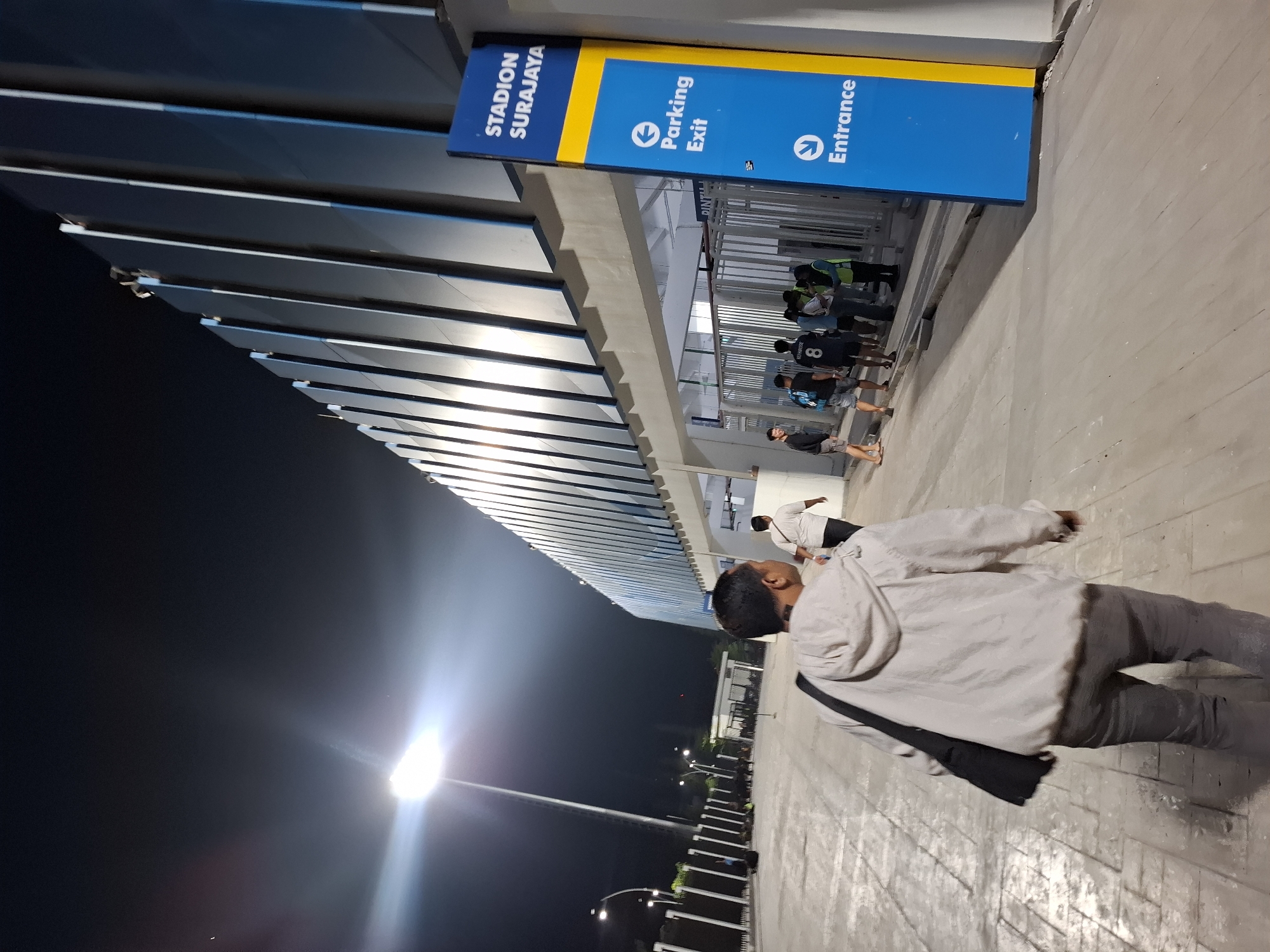 The image depicts the exterior of the Surajaya Stadium in Perumda Deket, Indonesia, at night. A person wearing a light-colored hooded jacket and dark pants walks away from the camera towards the stadium entrance. To the right, a tall blue and yellow sign indicates "STADION SURAJAYA" with arrows pointing to "Parking Exit" and "Entrance." Several people are gathered near the entrance, which is gated and lit by overhead lights. The stadium building features a modern design with vertical paneling. The ground is paved, and a bright stadium light illuminates the area.
