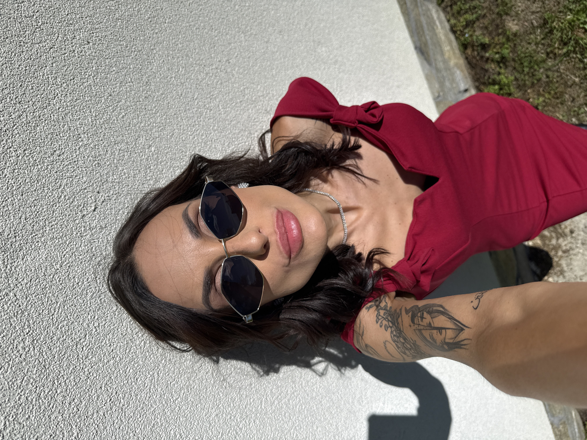 A woman in a red top and sunglasses is taking a selfie. The woman is positioned against a textured white wall. She has dark hair and is wearing a diamond necklace. Her arm is extended towards the camera, revealing a tattoo. The background includes a patch of grass and a gray concrete ledge. The location appears to be outdoors in a sunny setting, but Ribaritsa, Bulgaria, cannot be forensically confirmed.