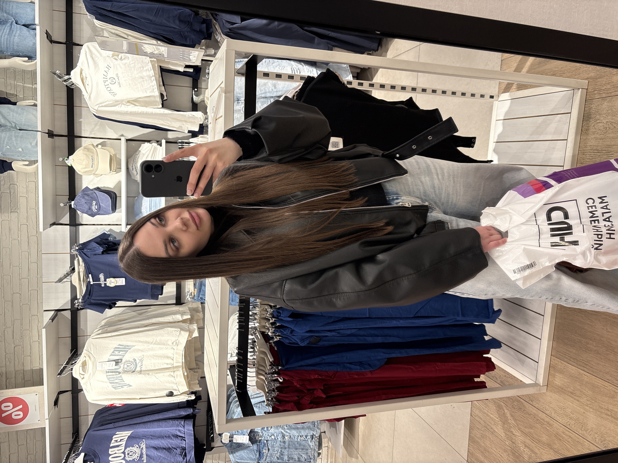 A young woman is captured in a mirror selfie inside a clothing store, likely located in Yekaterinburg, Russia. The image is oriented with the woman lying on her side. She has long, straight brown hair and is wearing a black leather jacket over a black turtleneck, paired with light-wash blue jeans. She holds a black smartphone (appears to be an iPhone) in her right hand, taking the photograph, and a white plastic shopping bag in her left hand.
The setting is a retail environment with white display shelves and racks filled with various garments, including blue, red, and cream-colored shirts, as well as denim jeans. Some shirts feature printed text, with "NEW YORK" and "AMERICAN" being partially visible and reversed. A red circular price tag showing a "%" symbol (indicating a discount) is visible on a shelf to the left. The store features a light brick-patterned wall texture and light wood-effect flooring.
The white shopping bag she is holding has reversed text, which appears to read "СПАР СЕМЕЙНЫЙ МАГНИТ" (SPAR SEMEYNY MAGNIT), indicating it's likely a supermarket bag rather than one from the clothing store. A barcode is also visible on the bag. Her expression is neutral as she looks towards the camera. The store appears well-lit, typical of a daytime or evening shopping experience.
