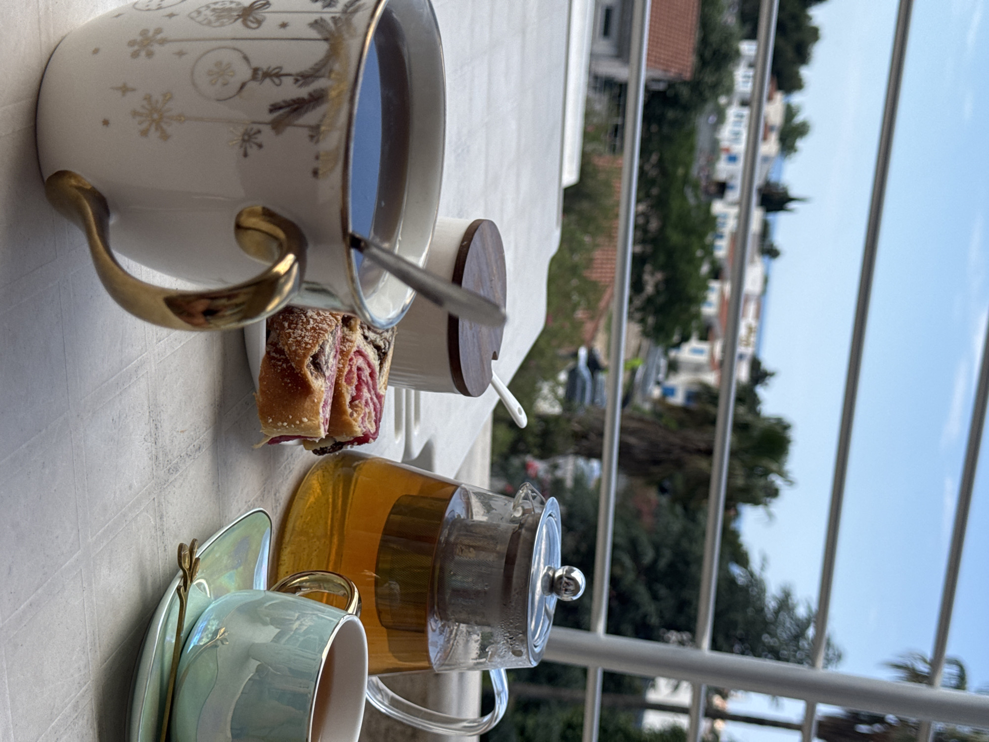 The image depicts a table set for tea on a balcony in Budva, Montenegro. A white teacup with gold accents sits next to a small pastry filled with red jam. A silver spoon rests in the cup. Behind the white teacup is a small white sugar bowl with a dark wooden lid and a white spoon sticking out. In the foreground, there is a pale green teacup on a matching saucer with a gold spoon. A clear teapot filled with yellow tea sits beside the green teacup. The balcony overlooks a cityscape with buildings, trees, and the sea in the distance. A white metal balcony railing is visible. The sky is bright blue.