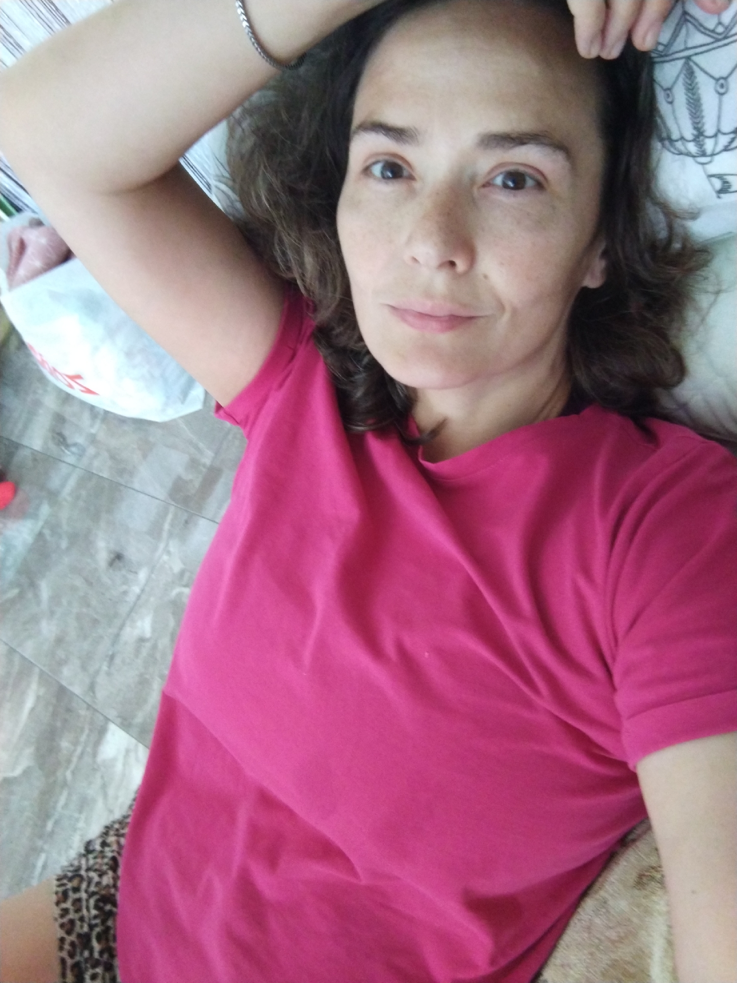 A woman is lying on her side in what appears to be a bedroom. She is wearing a bright pink t-shirt and leopard print shorts. Her left arm is bent above her head, and her right arm is resting on her leg. The woman has brown hair and freckles on her face. The background includes a bed with a patterned pillow and a floor that appears to be made of grey tiles. A white plastic bag is visible on the left side of the image. No specific details about the location in Vinogradny, Russia can be determined from the image alone.