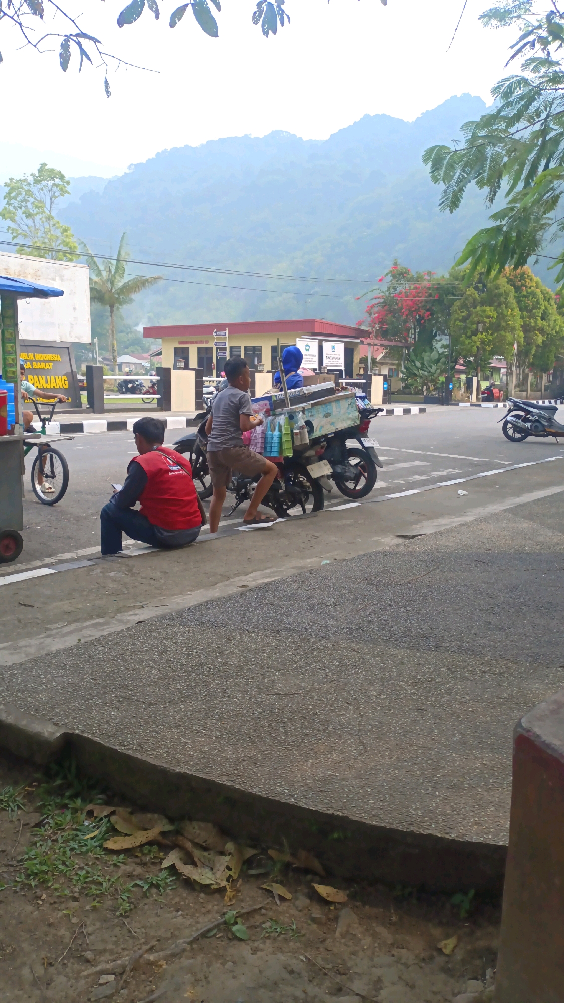 A slightly elevated, outdoor, daytime, vertical, medium shot shows a street scene in Padang Panjang, Indonesia, with a misty mountain range in the background. In the foreground, a young boy wearing a grey t-shirt and brown shorts is sitting on the back of a parked motorcycle with a blue covered cart attached. He is holding a small white food item and looking towards the left. Next to him, a person wearing a blue headscarf is also on the motorcycle. To the left of the motorcycle, a young man wearing a red vest over a dark long-sleeved shirt and blue pants is sitting on the curb, looking down at his phone. A small red and white cart is partially visible to the far left. In the middle ground, a building with a red roof and beige walls is visible, with a sign that reads "POLISI" and "REPUBLIK INDONESIA". Several parked motorcycles are also present. The street is paved with asphalt and has white and black striped markings. The background features a lush green mountain range covered in mist. Trees and foliage are visible on the right side of the image.