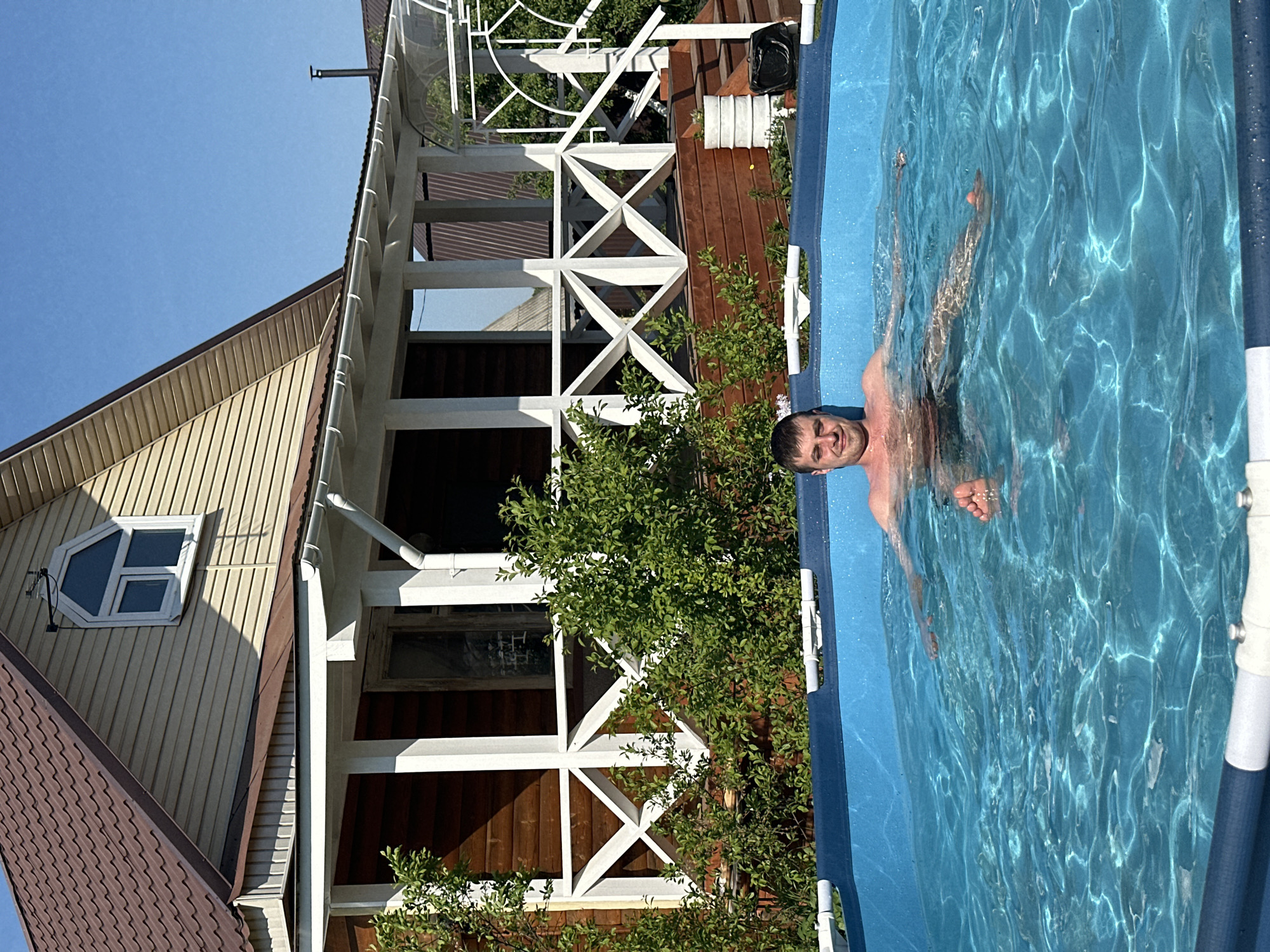 A man swims in an above-ground pool next to a wooden house in Khokhryaki, Russia. The house has brown siding and a white porch. The sky is blue.
