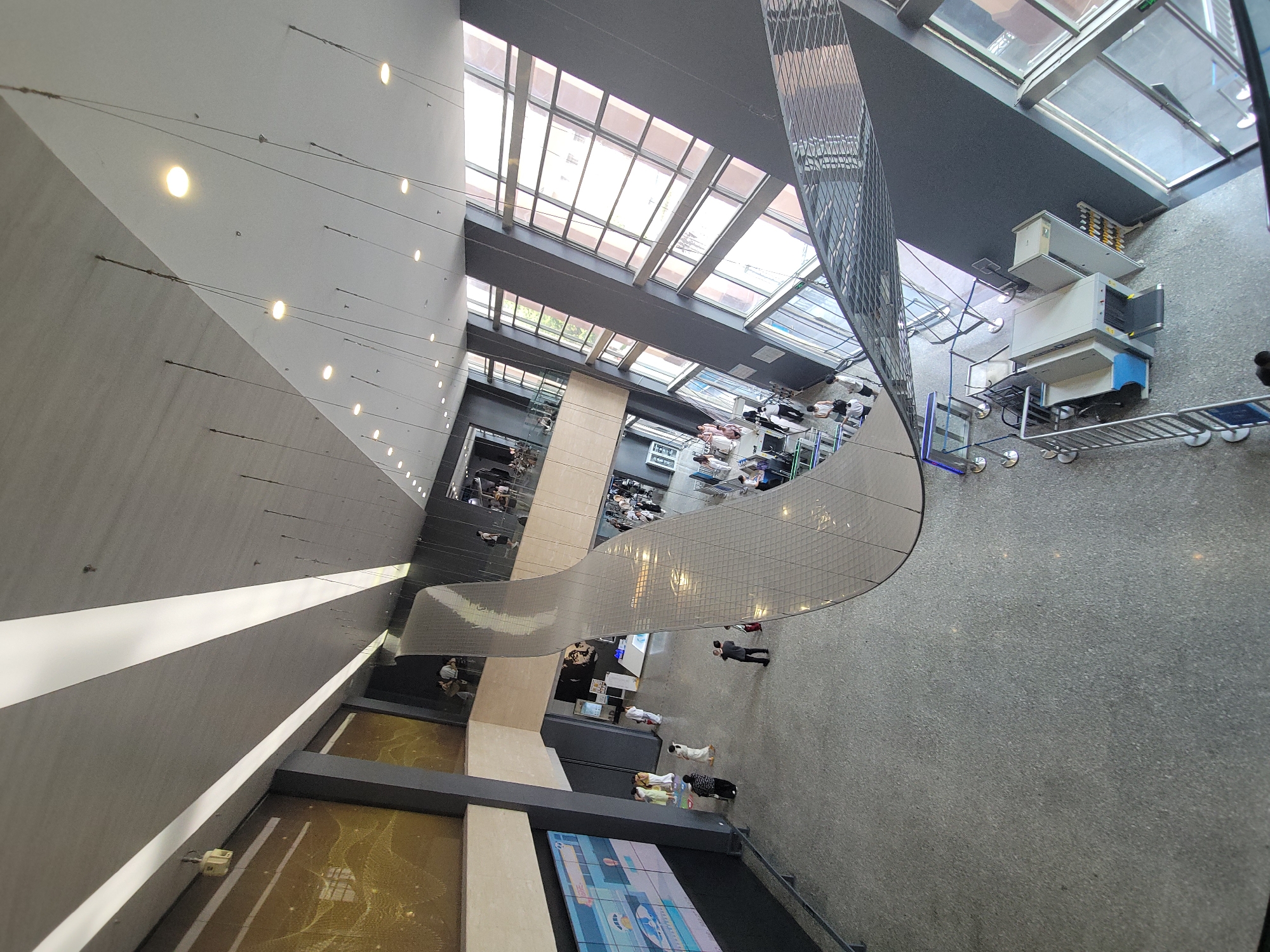 The image shows a wide, multi-level atrium with a large, undulating metallic sculpture suspended from the ceiling. The sculpture is composed of many small, reflective rectangular panels. The atrium features a grey, speckled floor, dark grey walls, and a grey ceiling with linear LED lighting. Large windows on the right side of the image offer views of an urban environment, with visible trees and buildings.
On the ground floor, several individuals are present. Two people in white clothing stand near a reception area on the left, next to a large digital screen displaying a colorful graphic. Further into the atrium, a man in a dark suit stands, facing away from the camera, holding a red object. A metal detector and security barriers are visible in the lower right section of the image, indicating a security checkpoint.
On an upper level, also visible in the atrium, are more people gathered, some seated and some standing, appearing to interact or observe the space. The overall impression is of a modern, spacious public building, possibly an exhibition hall, museum, or corporate lobby. The specific location in Qingyang District, China is not discernible from the visual information provided.