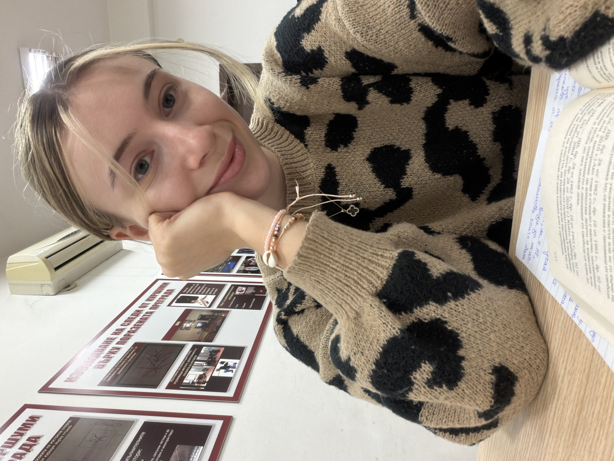 A fair-skinned young woman with blonde highlighted hair rests her chin on her hand. She is wearing a beige and black leopard print sweater. She is sitting at a table with an open book in front of her. On the table are printed displays with photographic and diagrammatic content on ballistics analysis, which include sections in Bulgarian Cyrillic script.
There is a unit of air conditioning equipment above the displays on the wall. The location can be inferred as Plovdiv, Bulgaria from the Cyrillic text.