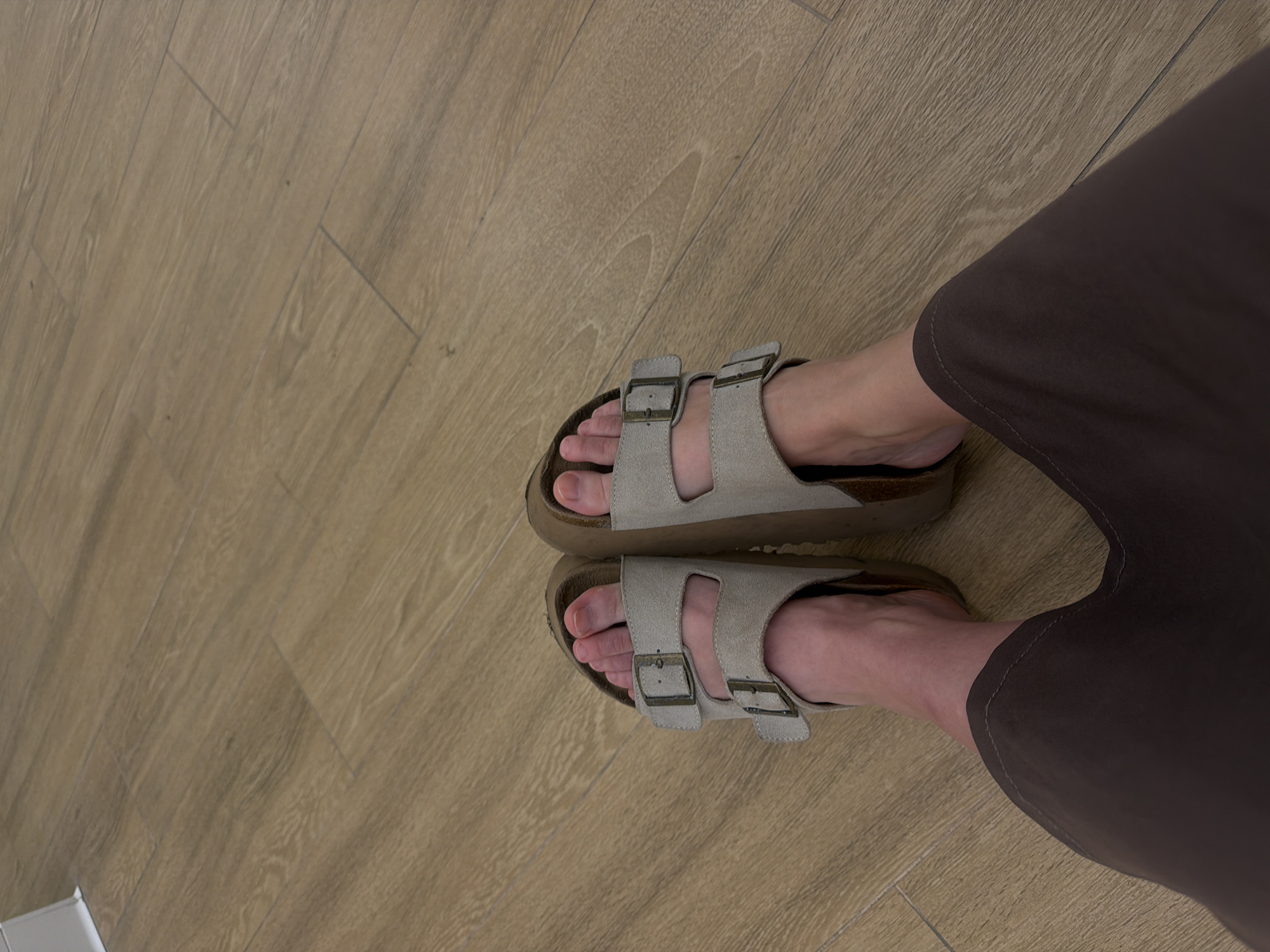 The image shows a pair of feet wearing Birkenstock Arizona sandals on a light brown wood-look floor. The person is wearing dark brown pants. The location is not discernible from the image, so the city of Moscow cannot be confirmed.