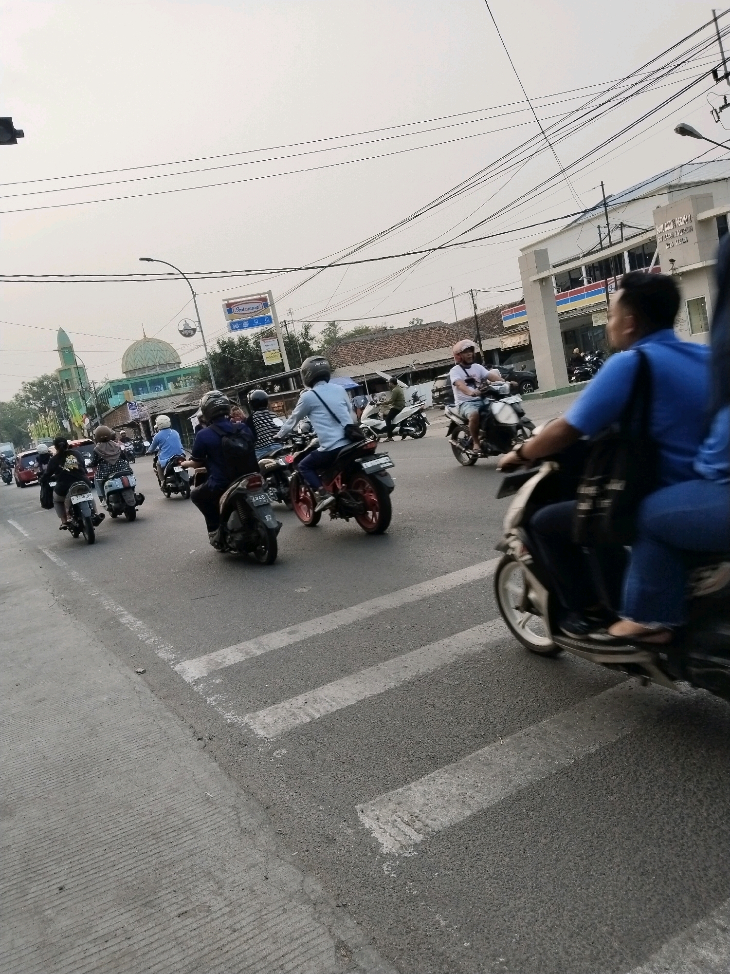 The image captures a street scene in Serang, Indonesia, focusing on a busy intersection with multiple motorcycles in motion. A prominent green-domed building, likely a mosque, is visible in the background, along with commercial establishments. Several individuals on motorcycles are navigating the road, some are wearing helmets, and others are not. The foreground shows a pedestrian crossing with white stripes on the asphalt. The overall environment appears to be an urban setting with overhead wires and buildings lining the street. The lighting suggests it is daytime, possibly late afternoon, with an overcast sky.