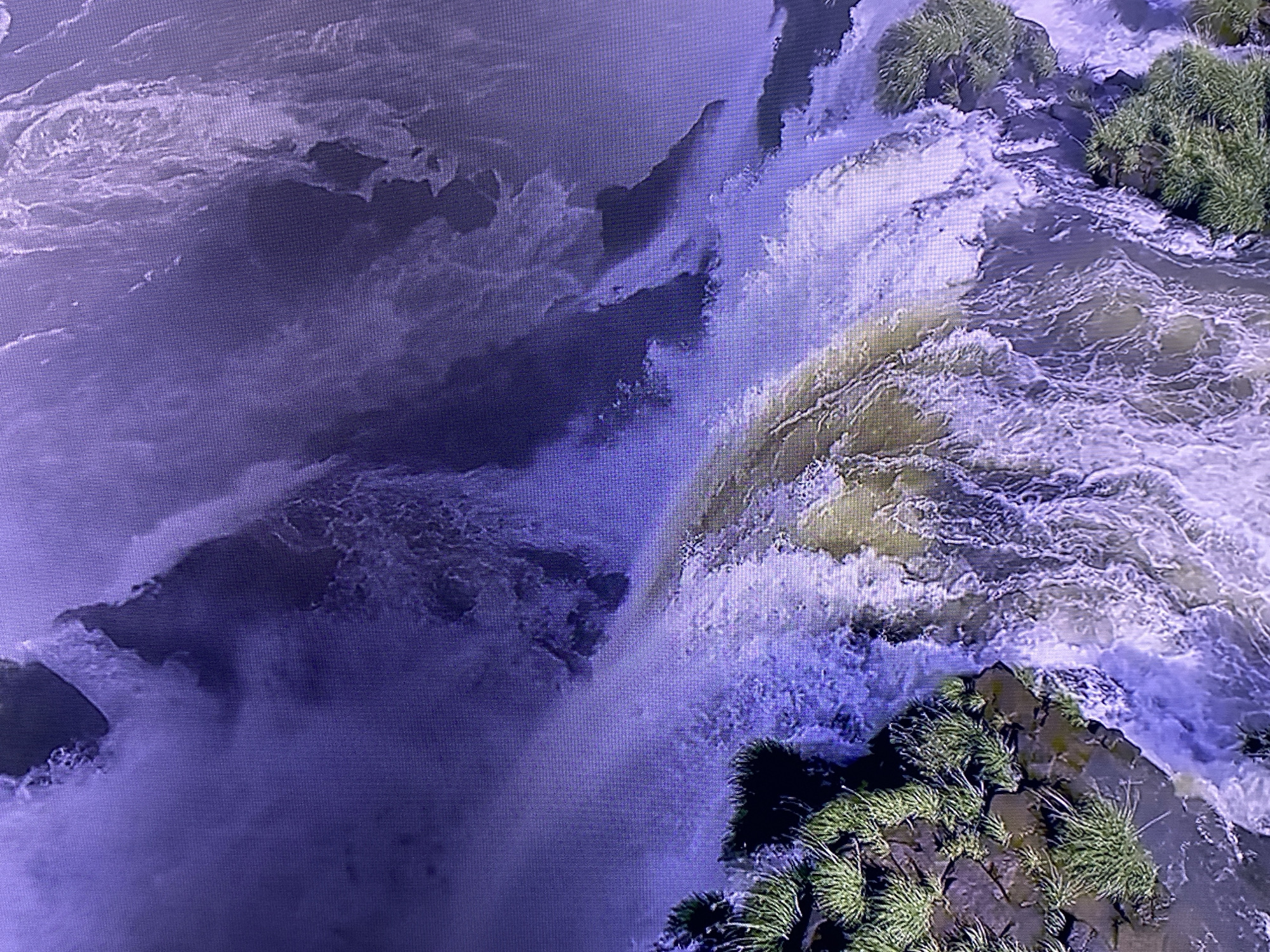 This media file depicts an aerial view of a powerful waterfall. The primary subjects are the cascading water and the surrounding rocky landscape with patches of dense green vegetation. The water is a mix of white foam and darker, sediment-laden currents, suggesting a forceful flow. The rocks are dark and appear wet, with some jutting out into the water. The vegetation consists of lush green grasses and possibly small bushes, clinging to the rocky edges.
The activity taking place is the natural phenomenon of a waterfall in motion, with water plunging over a precipice and churning below. The scene is dynamic and suggests a natural, untamed environment.
Notable details include the dramatic contrast between the bright white spray of the waterfall and the darker, more muted tones of the water and rocks. The vegetation adds a touch of life and color to the otherwise rugged landscape. The overall impression is one of immense natural power and beauty. There are no visible people, objects other than natural elements, visible text, or clear indicators of time of day or specific weather conditions, although the water's movement implies it is likely not frozen. The location is not explicitly identified.