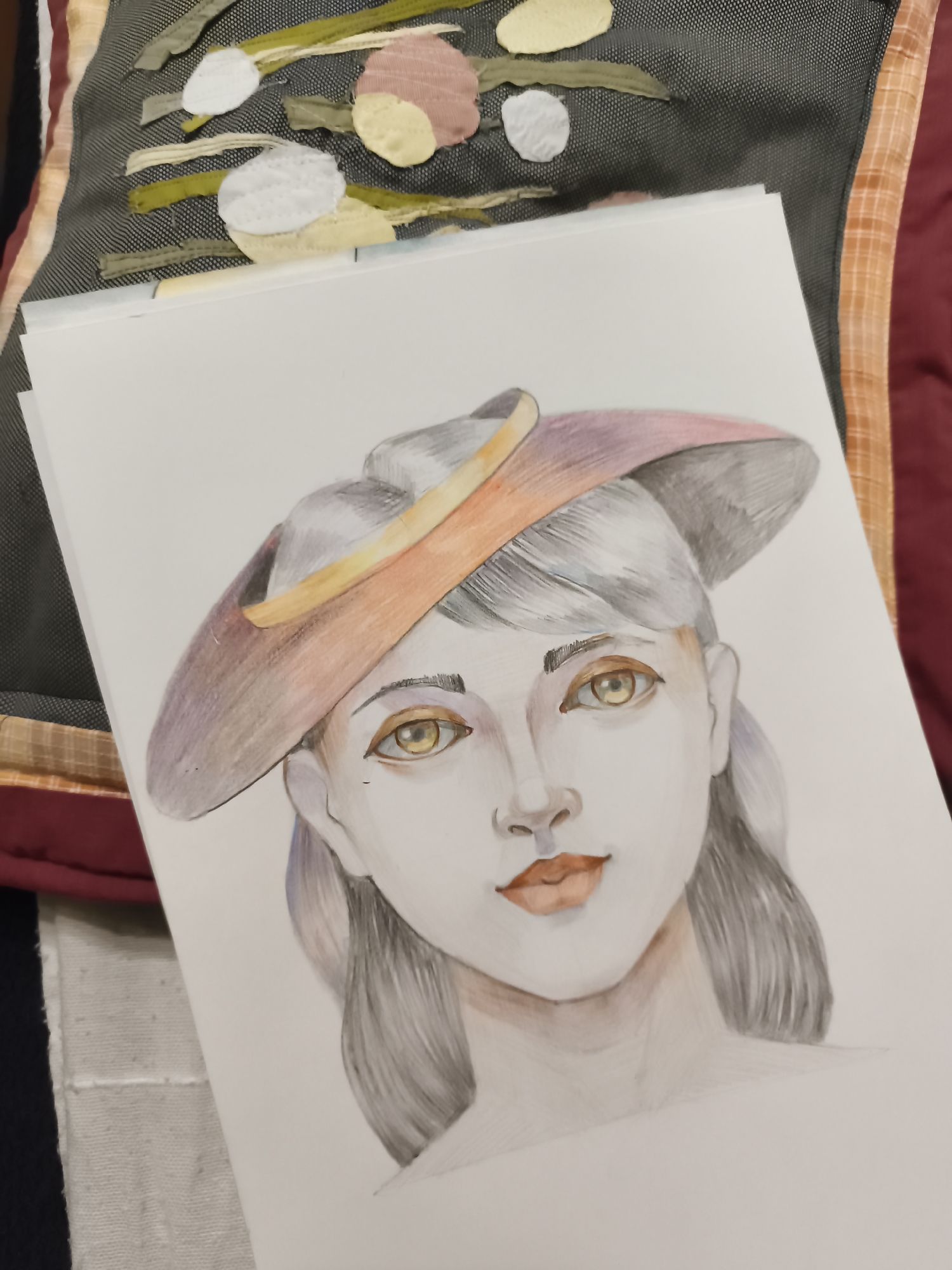 The image displays a watercolor painting of a woman's face, seen from the chest up, wearing a wide-brimmed hat. The woman has fair skin, gray hair, and striking yellow eyes. Her lips are painted a reddish-orange color. The hat is depicted with shades of purple, pink, and orange. The painting is on a white sheet of paper, which is placed on top of other papers. In the background, there is a textile surface with a pattern of abstract shapes in various colors, including green, yellow, pink, and white. This textile appears to be part of a decorative element, possibly a cushion or wall hanging. The overall scene is likely an indoor setting, and there is no indication of the city of Saint-Denis, France.
