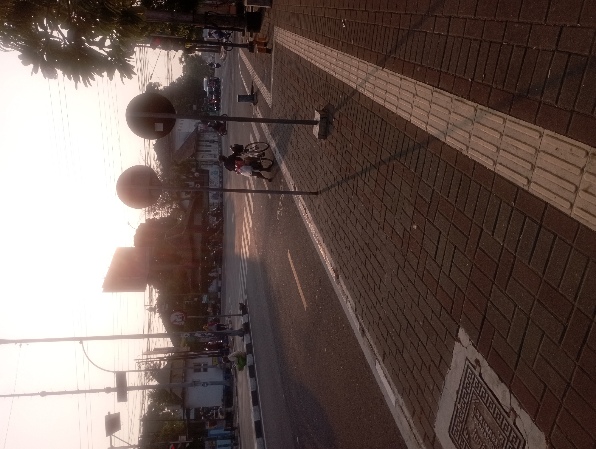 The image depicts an outdoor scene in Yogyakarta, Indonesia, likely near an intersection, during a sunny day with low-angle sunlight casting long shadows. The foreground is dominated by a brick-paved sidewalk with tactile paving. To the left of the sidewalk is a road with a pedestrian crossing. In the middle ground, two large circular traffic signs on poles are visible, one slightly obscured by the other. A person is pushing a bicycle laden with bags across the pedestrian crossing. A few other individuals are seen on the sidewalk and in the distance, some of whom appear to be on motorcycles. Several buildings are present in the background, as are utility poles with numerous wires and some trees. A traffic light is visible on the right side of the frame. The overall impression is of a moderately busy urban environment at what appears to be late afternoon or early morning due to the lighting.