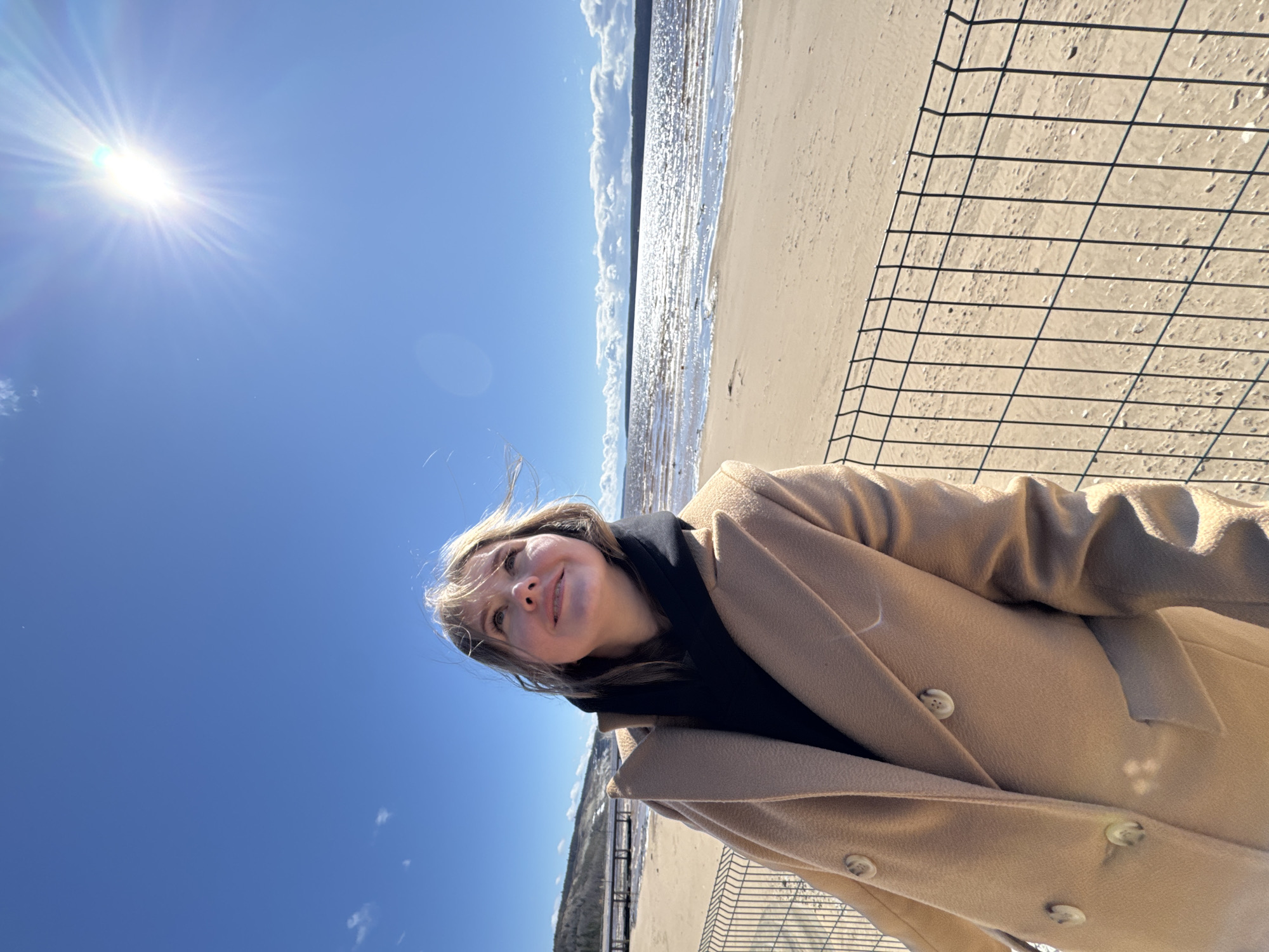 This slightly tilted, low-angle selfie captures a young woman outdoors under a bright, sunny sky in Polazna, Russia. The woman, likely in her 20s or 30s, is positioned in the lower mid-right, wearing a light brown or camel-colored long coat with prominent buttons and a black scarf or turtleneck. Her light-colored hair is softly blowing, and she displays a gentle, pleasant smile while looking slightly upwards and away from the camera.
The scene depicts a clear, sunny day, with the bright sun flaring intensely in the upper left corner of the deep blue sky. The background shows a wide sandy beach or shore meeting a body of water, likely the Kama Reservoir, which reflects the sunlight in shimmering patterns. Across the water, a distant, faint shoreline with some trees or land features is visible. A black wire fence or grid diagonally crosses the lower right foreground, partially obscuring the sandy surface. The presence of the coat suggests it might be cool despite the bright sunshine. There is no visible text in the image.