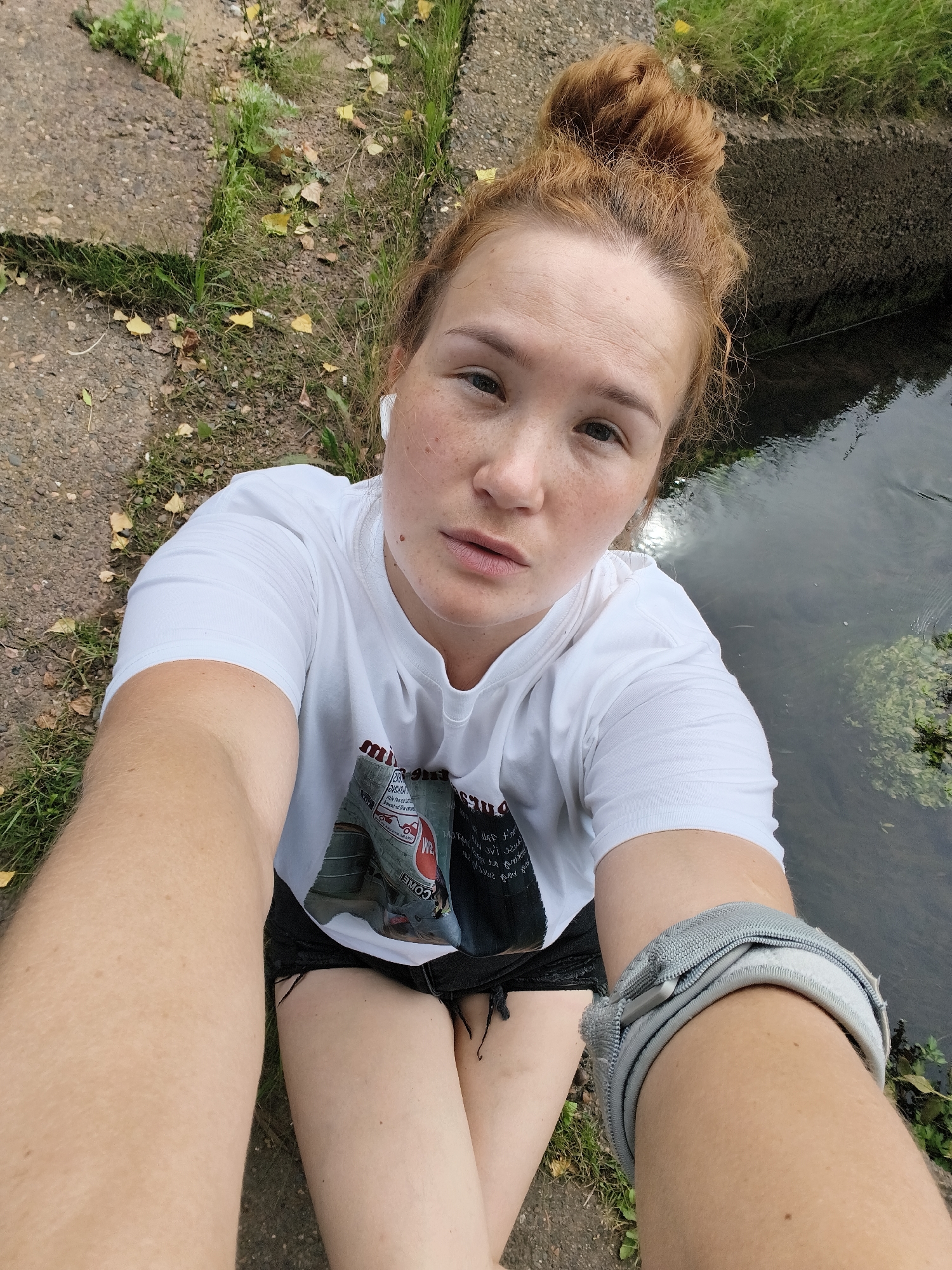 A young woman with red hair tied in a bun is taking a selfie. She is wearing a white t-shirt with a graphic print on the front, black shorts, and white earbuds. A grey band is visible on her right arm. She has freckles across her nose and cheeks. She is sitting on the ground next to a body of water, possibly a river or canal. The ground is a mix of concrete slabs, dirt, and grass with some fallen leaves. The water appears murky and there is some green vegetation growing in it. The lighting suggests it is daytime, possibly overcast due to the lack of harsh shadows. The woman's expression is neutral, and she is looking directly at the camera. The overall scene conveys a casual, outdoor setting. There is some text visible on the graphic print of her t-shirt, but it is mostly obscured and illegible. The crops show a closer view of the woman's face, hair, and arm, as well as the surrounding environment.
