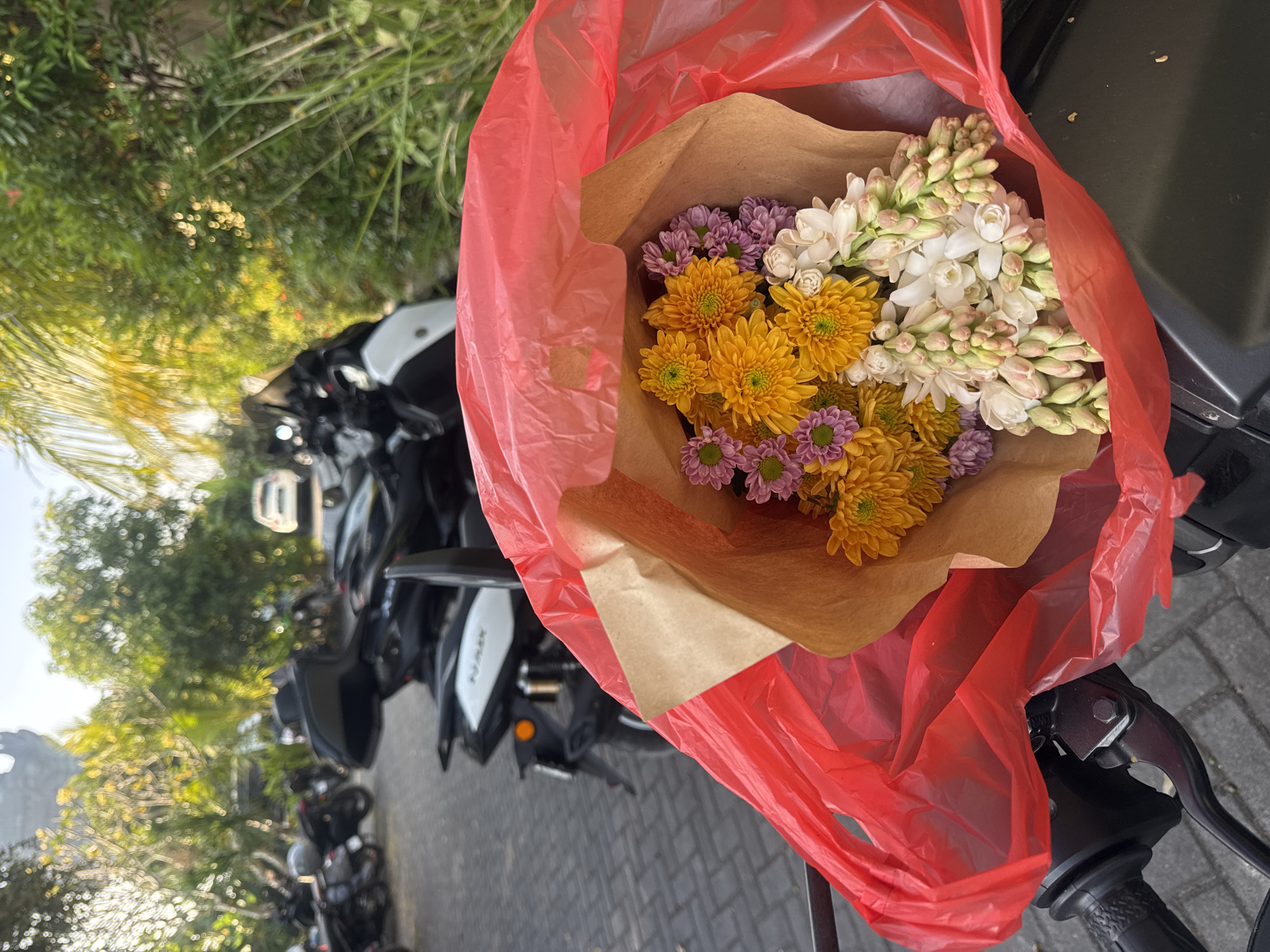 In Kuta Selatan, Indonesia, a bouquet of yellow chrysanthemums, purple flowers, and white tuberose is partially visible, wrapped in brown paper and a red plastic bag. The bouquet rests on a dark object, likely the handlebars of a motorcycle, which is parked on a paved surface. In the background, a line of parked motorcycles is visible, along with lush green foliage and trees, suggesting an outdoor setting possibly near a street or parking area. The lighting indicates it is daytime.