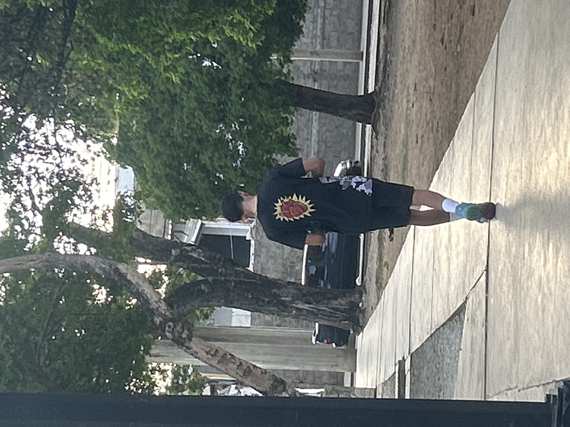 The image shows a person walking away from the camera on a sidewalk in Santa Ana, Venezuela. The person is wearing a black t-shirt with a graphic design of a heart with rays, black shorts, white socks, and blue and green athletic shoes. The person also appears to be wearing a dark baseball cap. The sidewalk is made of concrete slabs and is lined with trees and a low stone wall. In the background, there are buildings, a car, and more trees. The lighting suggests it is daytime, possibly late afternoon due to the angle of the light.