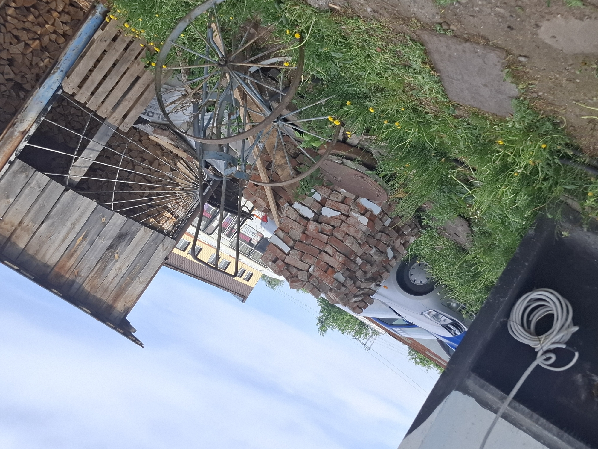 The image shows an outdoor scene, likely in a yard or storage area. There's a jumble of objects including two antique-looking metal wheels with spokes, a pile of red bricks, wood pallets, firewood stacked in a metal structure, and overgrown grass with yellow flowers. A white car is visible in the background, partially obscured by the bricks. The sky appears light blue with some clouds. The perspective is taken from a slightly elevated position, looking down at the objects. The overall scene gives the impression of a cluttered or disorganized outdoor storage space. The location in the city of Chaikovsky, Russia cannot be determined from the image.
