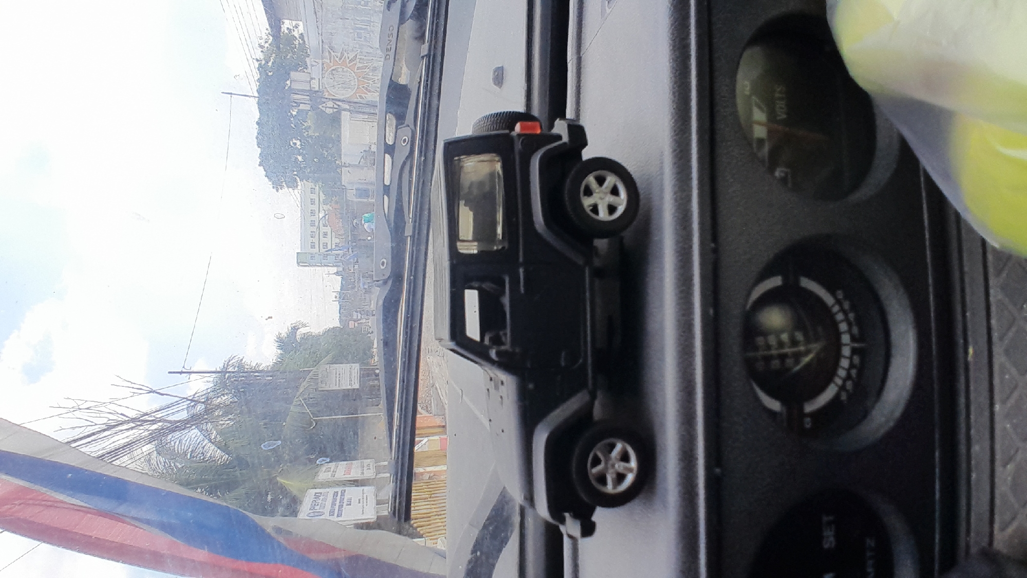 The image shows a close-up view of a black toy Jeep Wrangler parked on the dashboard of a vehicle. The toy Jeep is positioned in the foreground, with its front wheels and hood visible. Beyond the toy Jeep, the windshield of the vehicle reveals a blurred outdoor scene. This outdoor scene includes a street with trees, buildings, and a cloudy sky. A portion of a red, white, and blue flag is visible in the upper left corner of the image, attached to the interior of the vehicle. The dashboard also shows two gauges, one labeled "SET QUARTZ" and another with a needle pointing to "VOLTS". The scene appears to be an interior view from a vehicle in an urban or semi-urban environment, possibly in Bangkle, Indonesia, given the context.
