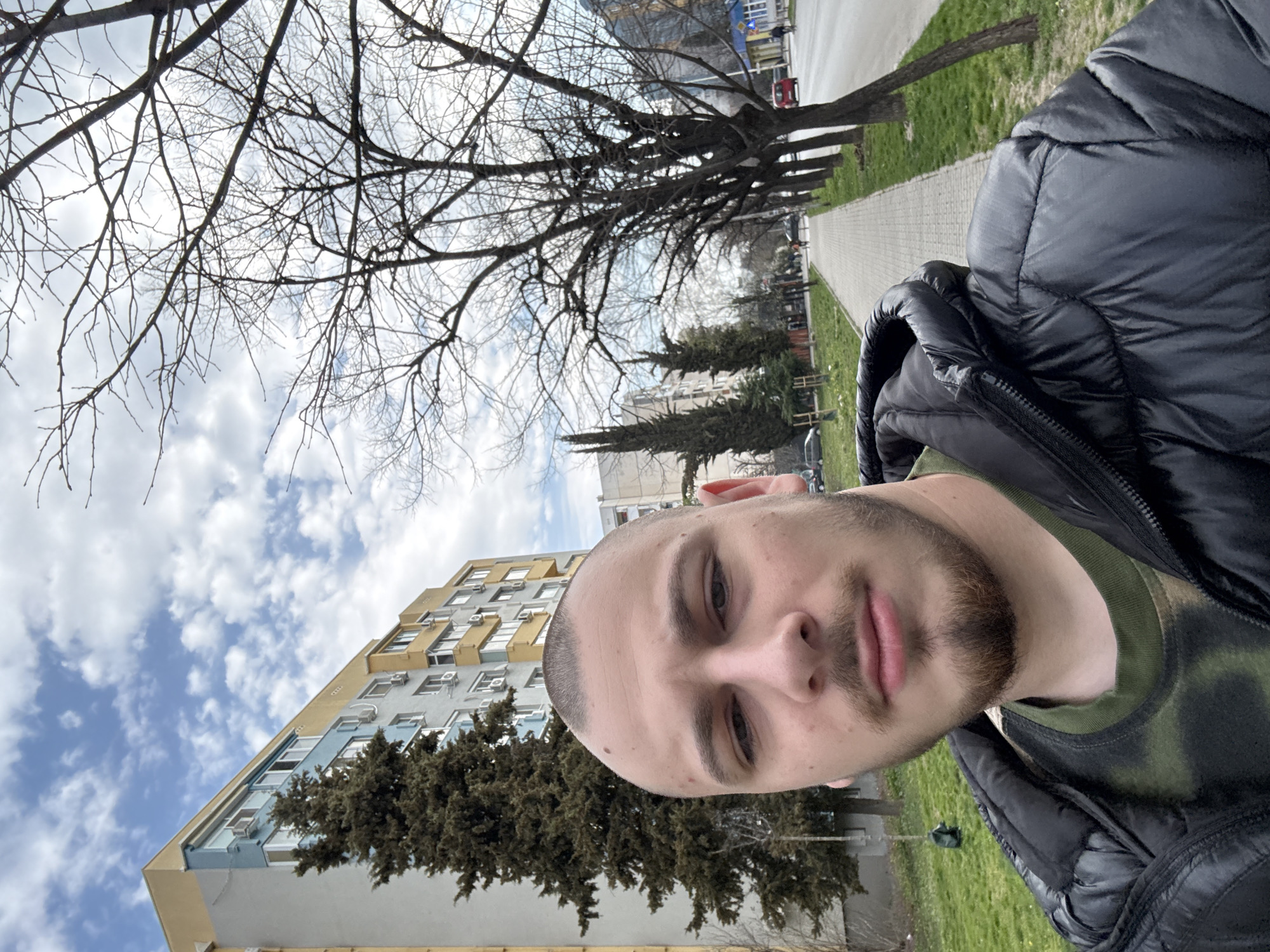 A male subject, approximately in his early twenties, with a shaved head, light brown beard, and mustache, takes a selfie. He is wearing a black quilted jacket over an olive green camouflage-patterned long-sleeved shirt. His expression is neutral.
The scene is outdoors in an urban environment within Stara Zagora, Bulgaria. In the background, to the left of the subject, is a multi-story apartment building with a light gray base, a light blue mid-section featuring numerous windows and external air conditioning units, and a yellow top section. A tall, dark green coniferous tree stands in front of this building. To the right, a paved sidewalk extends into the distance, bordered by a strip of green grass and a row of bare deciduous trees. Beyond the trees, an asphalt road is visible, with a red car and other vehicles in the distance. The sky is partially cloudy with blue patches. The presence of bare trees indicates late autumn, winter, or early spring. Several pedestrians are visible in the far distance walking along the sidewalk.