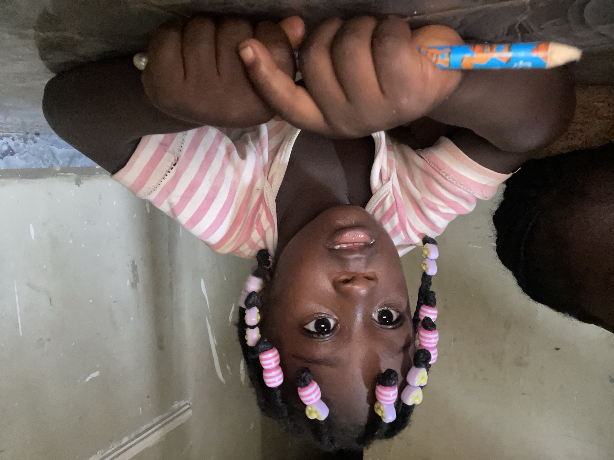 A young child, likely a girl with dark skin, is depicted in a head-and-shoulders portrait, looking directly at the camera with wide, dark eyes. She is holding a colorful blue and orange patterned pencil or crayon with both hands, which are clasped together near her chin. Her mouth is slightly open, and her expression appears curious and engaging.
The child wears a light-colored top with distinct pink and white horizontal stripes. Her black hair is styled in numerous braids, each adorned with a variety of colorful plastic beads in shades of pink, yellow, and purple, some featuring pink and white stripes.
The background suggests an indoor setting, possibly a room in Akwanga, Nigeria. The walls are a plain, light grayish-beige color and exhibit several scattered white paint splatters or marks, indicating a potentially utilitarian or lived-in space. The lighting is even, highlighting the child's features. The image captures a moment of childhood innocence, possibly during a creative or learning activity. No legible text is visible on the pencil or in the background.