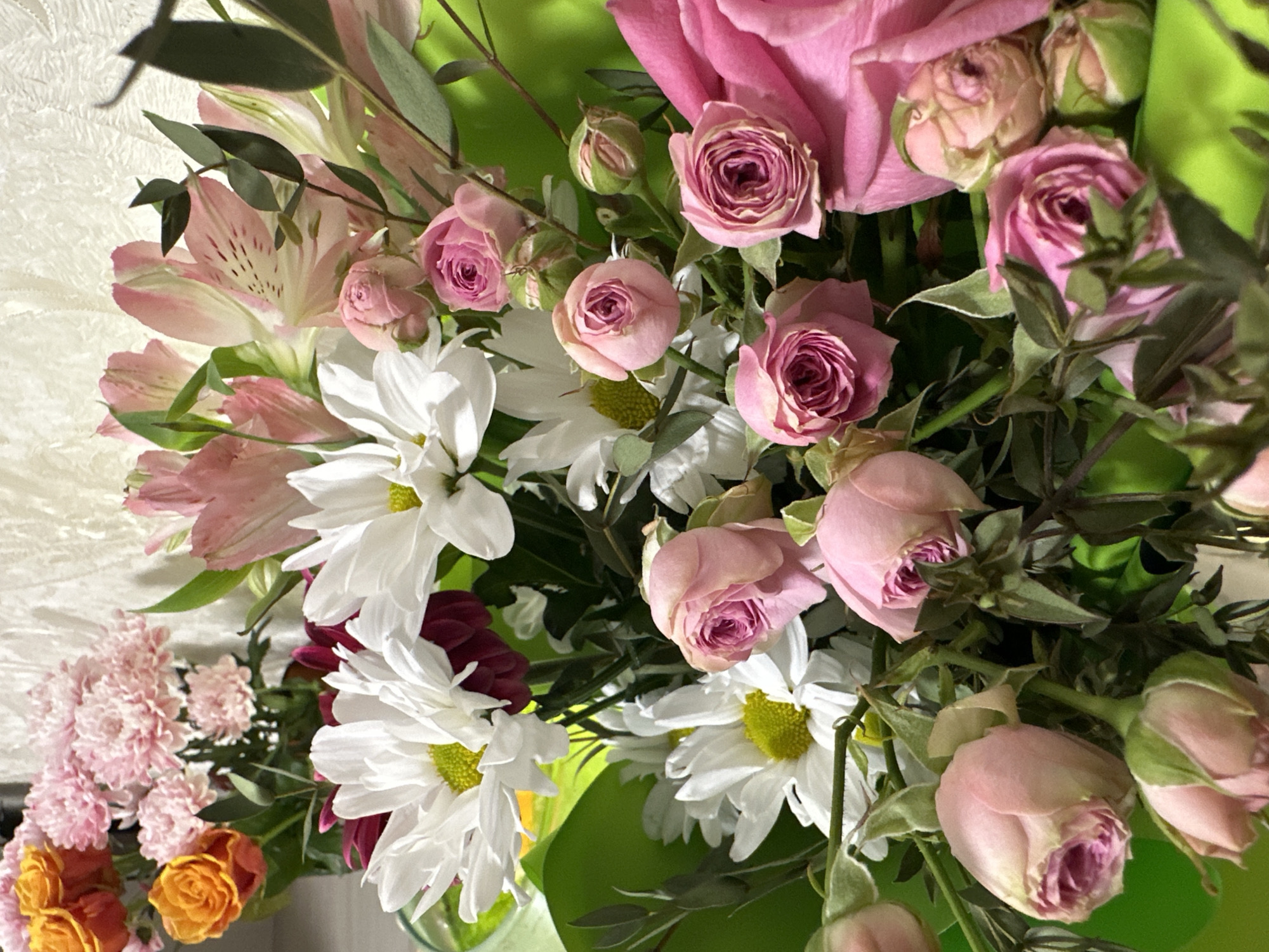 The image is a close-up shot of a bouquet of various flowers. Dominating the scene are pink roses, white daisies, and light pink alstroemeria. There are also smaller pink and orange flowers visible on the lower left. The background is a blurry green, likely foliage or wrapping paper of the bouquet. No people or specific locations are discernible in the image. The image suggests an indoor setting due to the controlled lighting and the nature of the subject. There is no evidence to suggest the location is in Uva, Russia.