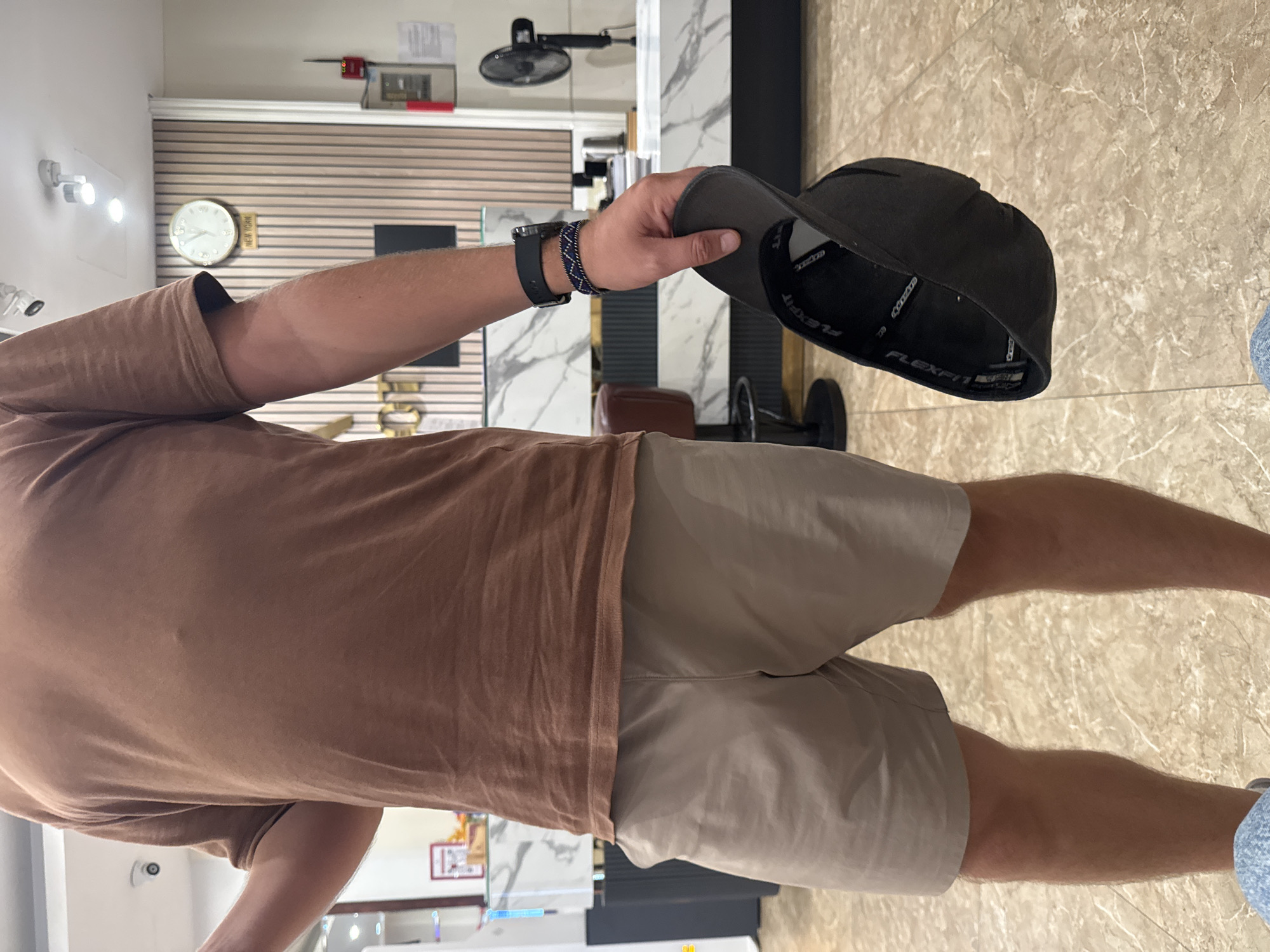 A person is standing in what appears to be an indoor setting, possibly a store or an office, given the presence of a clock on the wall and what looks like a reception desk in the background. The person is wearing a brown t-shirt and light beige shorts. They are holding a black baseball cap in their right hand. A black watch with a patterned band is visible on their left wrist. The floor is tiled with a marble-like pattern. In the background, there is a wall with horizontal wooden slats, a black oscillating fan mounted on the wall, and a clock displaying the time. There is also a sign that says "NEW YORK" next to the clock. The lighting suggests it is daytime. The person is facing away from the camera, and their posture does not convey any specific emotion. The overall scene is casual and domestic. The text "FLEXFIT" is visible on the inside of the baseball cap.