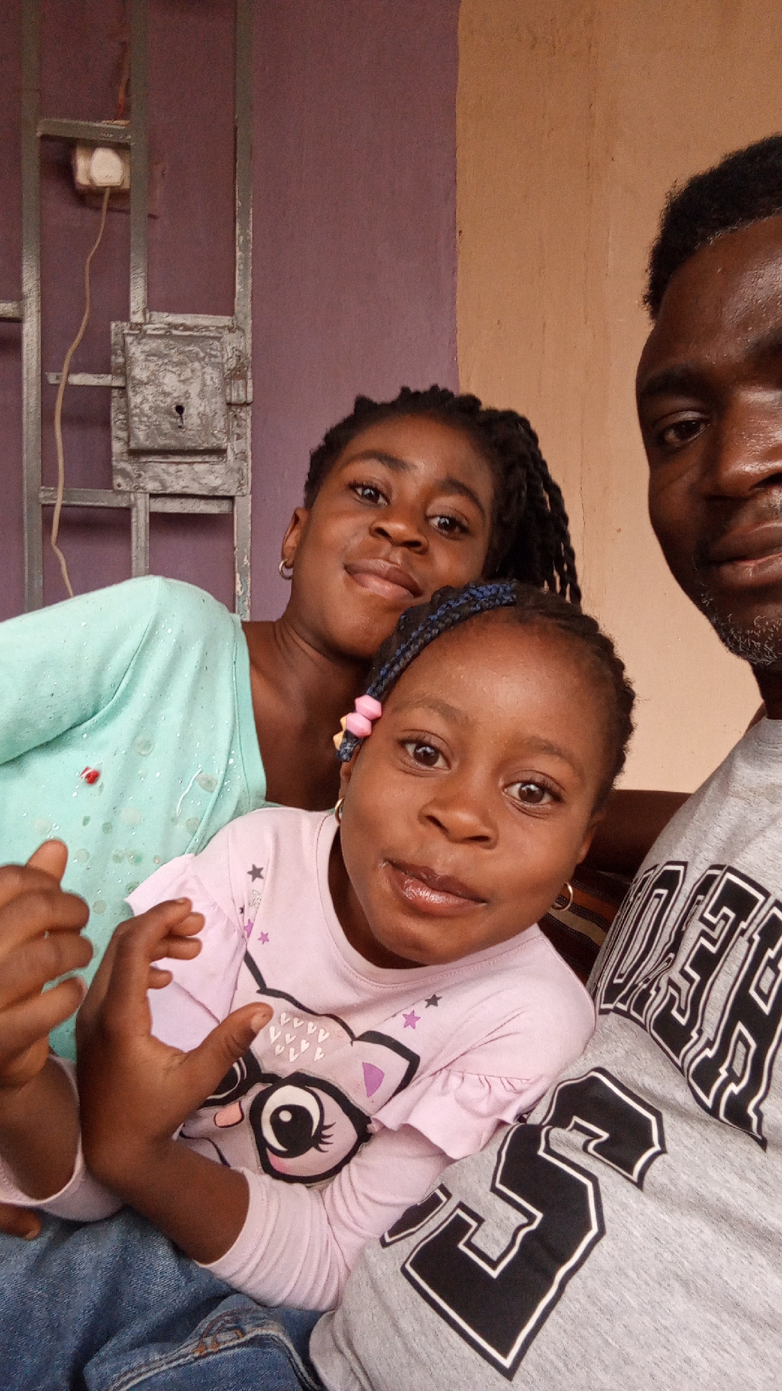 The image shows a selfie taken indoors, likely in a home. Three people are visible: two young girls and a man. One girl wears a light pink shirt with a cartoon animal on it, and the other wears a light green top. The man is wearing a grey shirt. The girls appear to be sitting close to the man. The background includes a purple wall with a metal gate or security door. There is an electrical outlet on the wall near the gate. Based on context, the location is likely Sabon Tasha, Nigeria.
