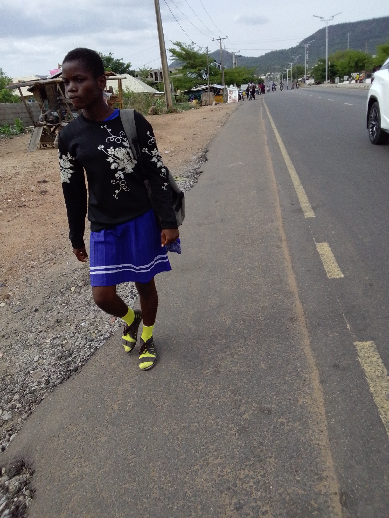 A person walks along the roadside in Adankolo, Nigeria. The individual, wearing a black long-sleeved shirt with white floral patterns, a blue skirt with white stripes, yellow socks, and sandals, carries a backpack. The scene includes a paved road with a yellow dividing line, a gravelly shoulder, and sparse buildings in the background. A hill is visible in the distance. Several people and vehicles are seen on the road further down the street. The sky is overcast. A white car is partially visible on the right side of the image.