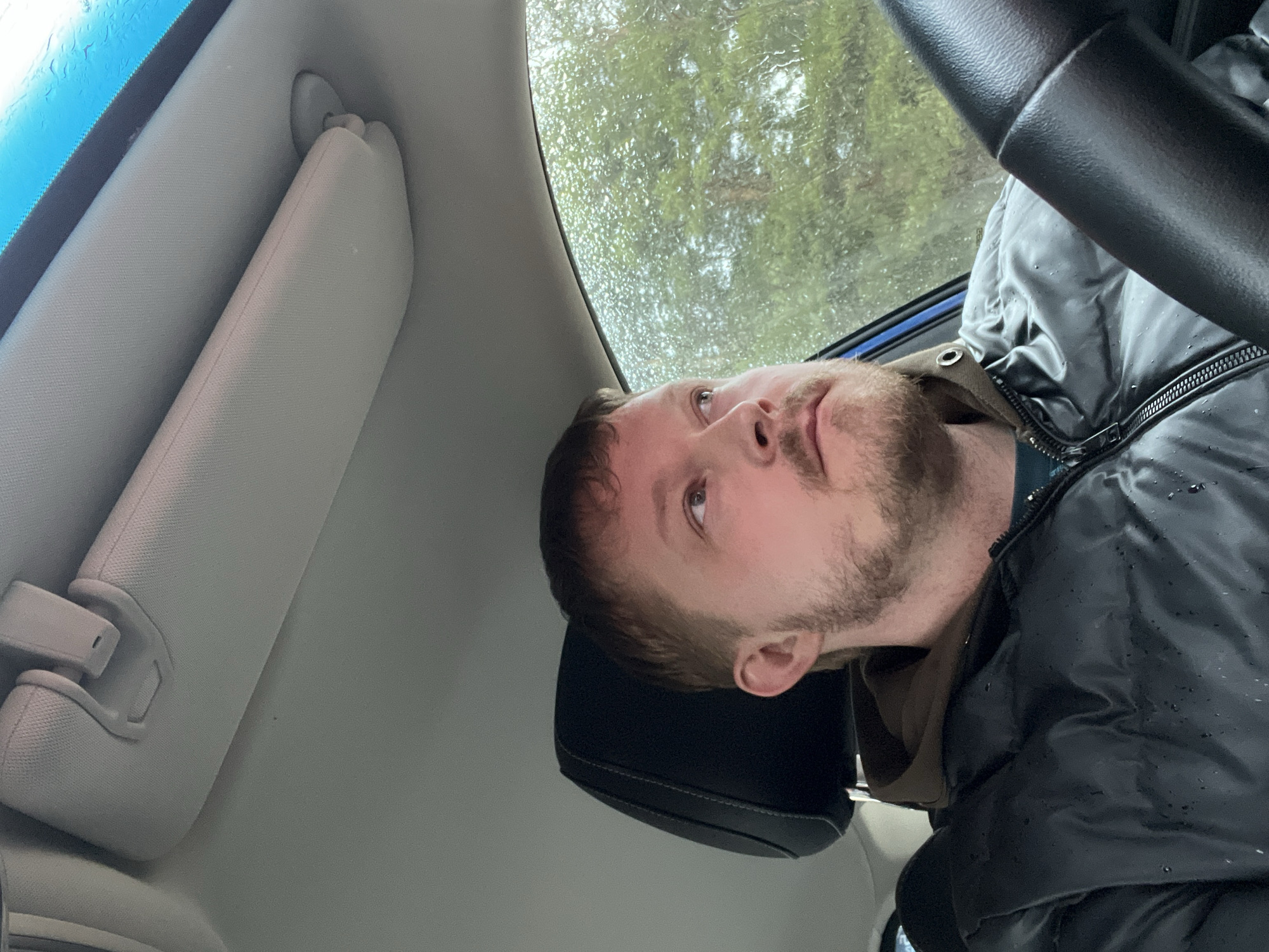 The image depicts a man sitting in the driver's seat of a car. Raindrops are visible on the windshield, suggesting it is raining. The car's sun visor is pulled down. The man is wearing a dark-colored jacket. The location in the city of Chaikovsky, Russia, is not determinable from the image.