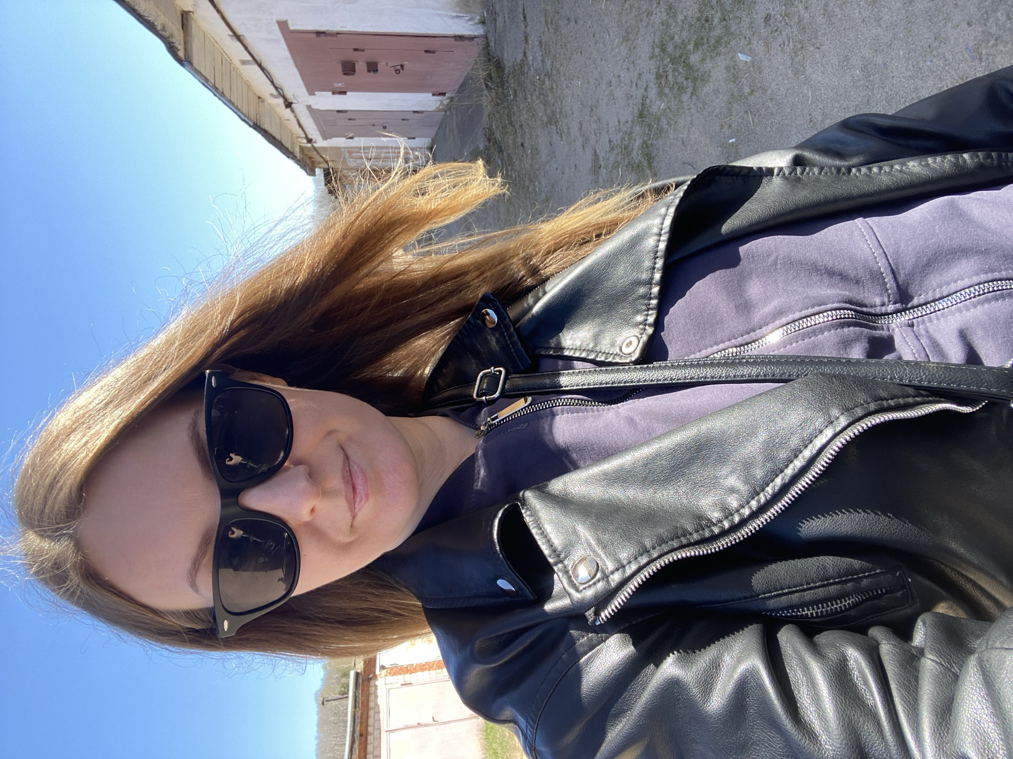 A woman in sunglasses and a black leather jacket takes a selfie outside a building, likely garages, in Noviy, Russia. The day is sunny. She is wearing a zip-up top under the jacket. The buildings are worn and of a light color.