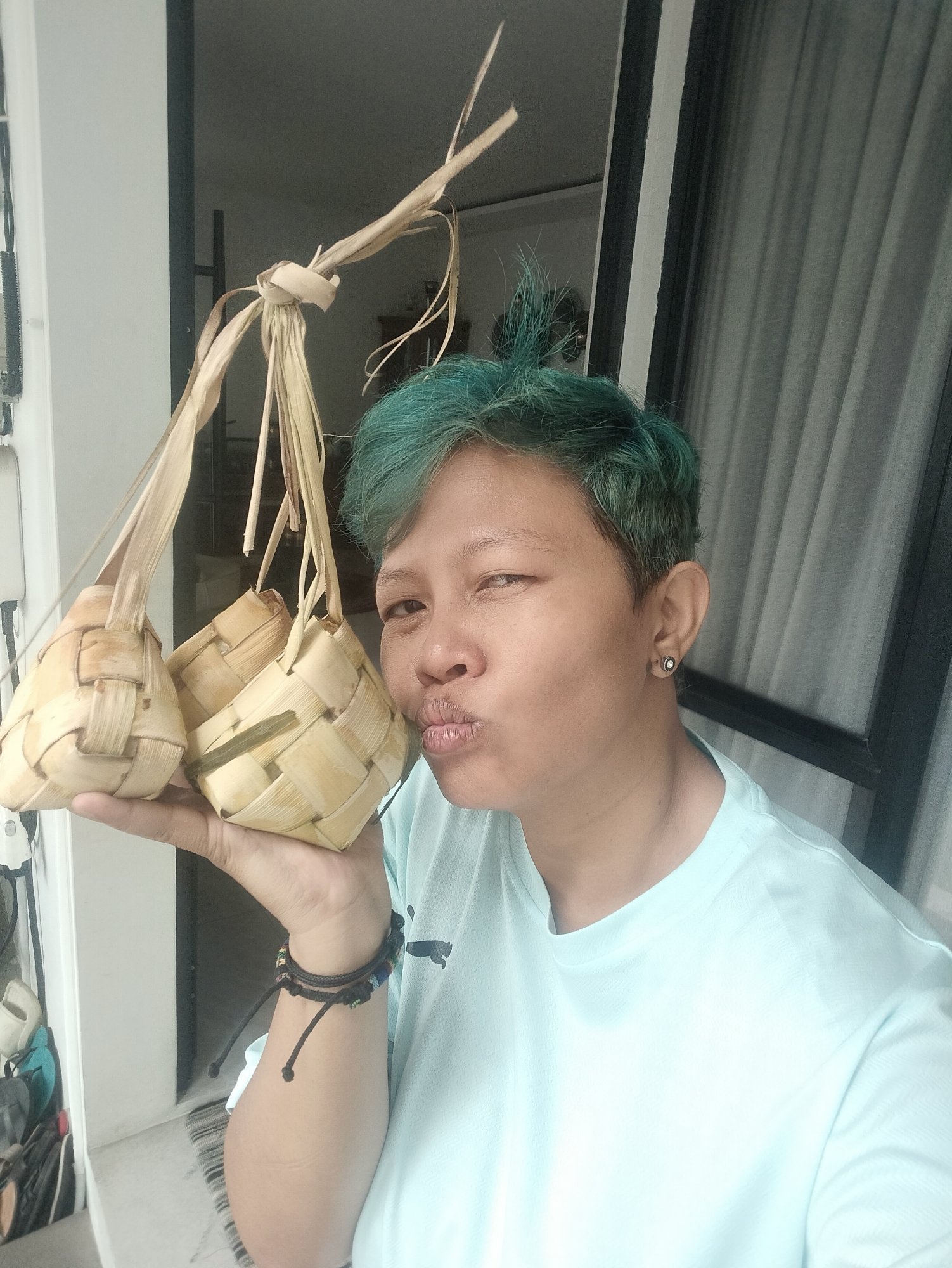 A person with short, dyed turquoise hair is holding three ketupat in their left hand, puckering their lips, possibly in a kissing gesture. The ketupat are woven from palm leaves and tied together with dried strips of the same material. The person is wearing a light blue t-shirt and a black bracelet on their left wrist. They also have a small earring in their left ear. They appear to be standing in front of a sliding glass door that is partially obscured by a curtain. The location is identified as Bekasi, Indonesia.