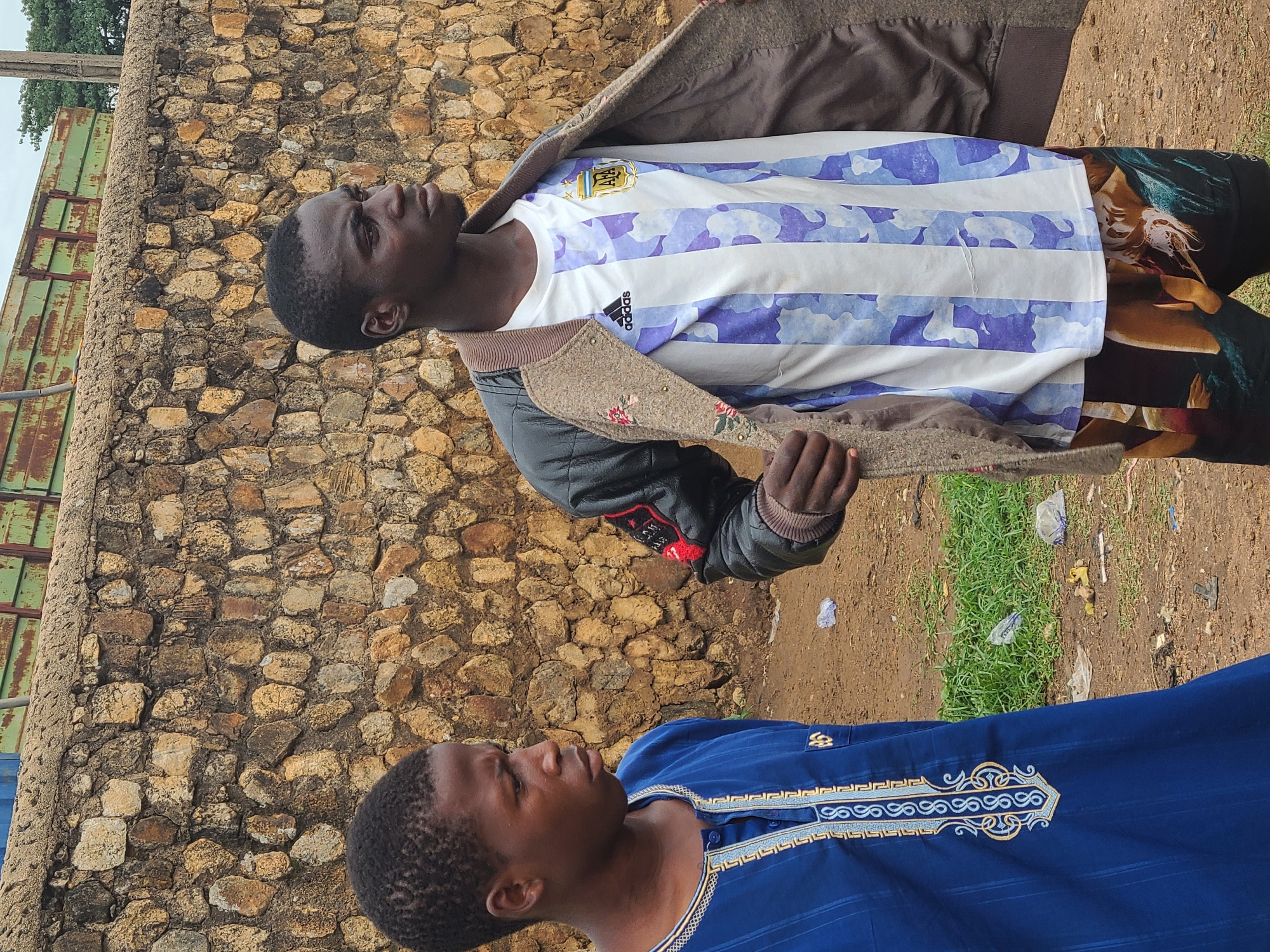 Two men are positioned outdoors with a stone wall in the background. The man on the upper left is wearing a white and blue horizontally striped shirt with the Argentina national football team crest, a brown jacket, and patterned pants. The man below is wearing a blue, patterned shirt. The ground is a mix of dirt and grass with litter scattered around.