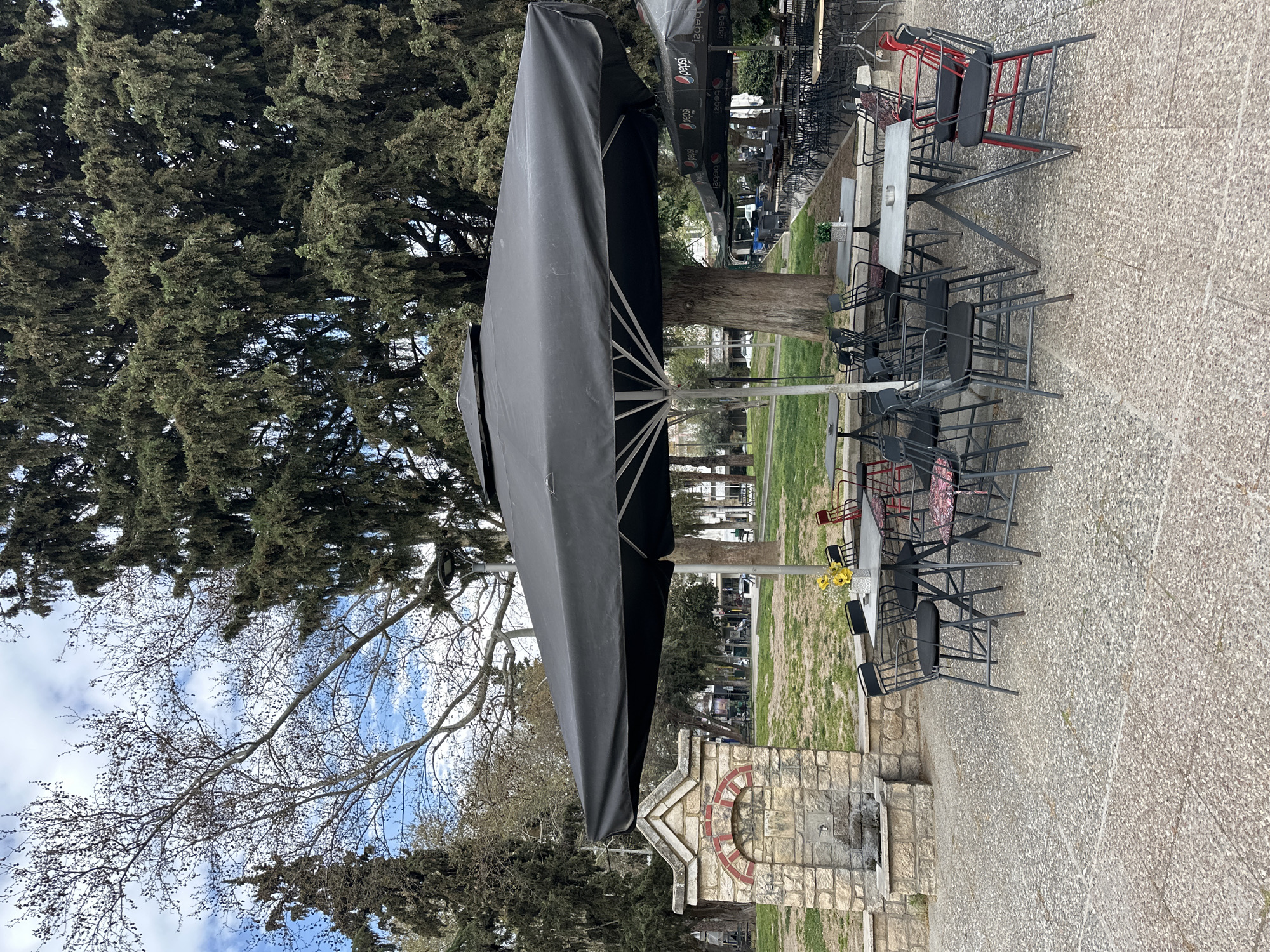 The image depicts an outdoor seating area of a cafe or restaurant in Serres, Greece. The tables and chairs are stacked or pushed together, suggesting the establishment is currently closed or preparing for closure. A large, closed black umbrella dominates the scene, casting a shadow over the adjacent tables. The tables are gray, while the chairs are mostly black with some red accents. The area is paved, and a low stone wall separates the seating from a grassy area. In the background, trees are visible. A stone fountain with a circular brick design is located near the seating. There is a vase of yellow flowers on one of the tables. The sky is visible through the bare branches of a tree.