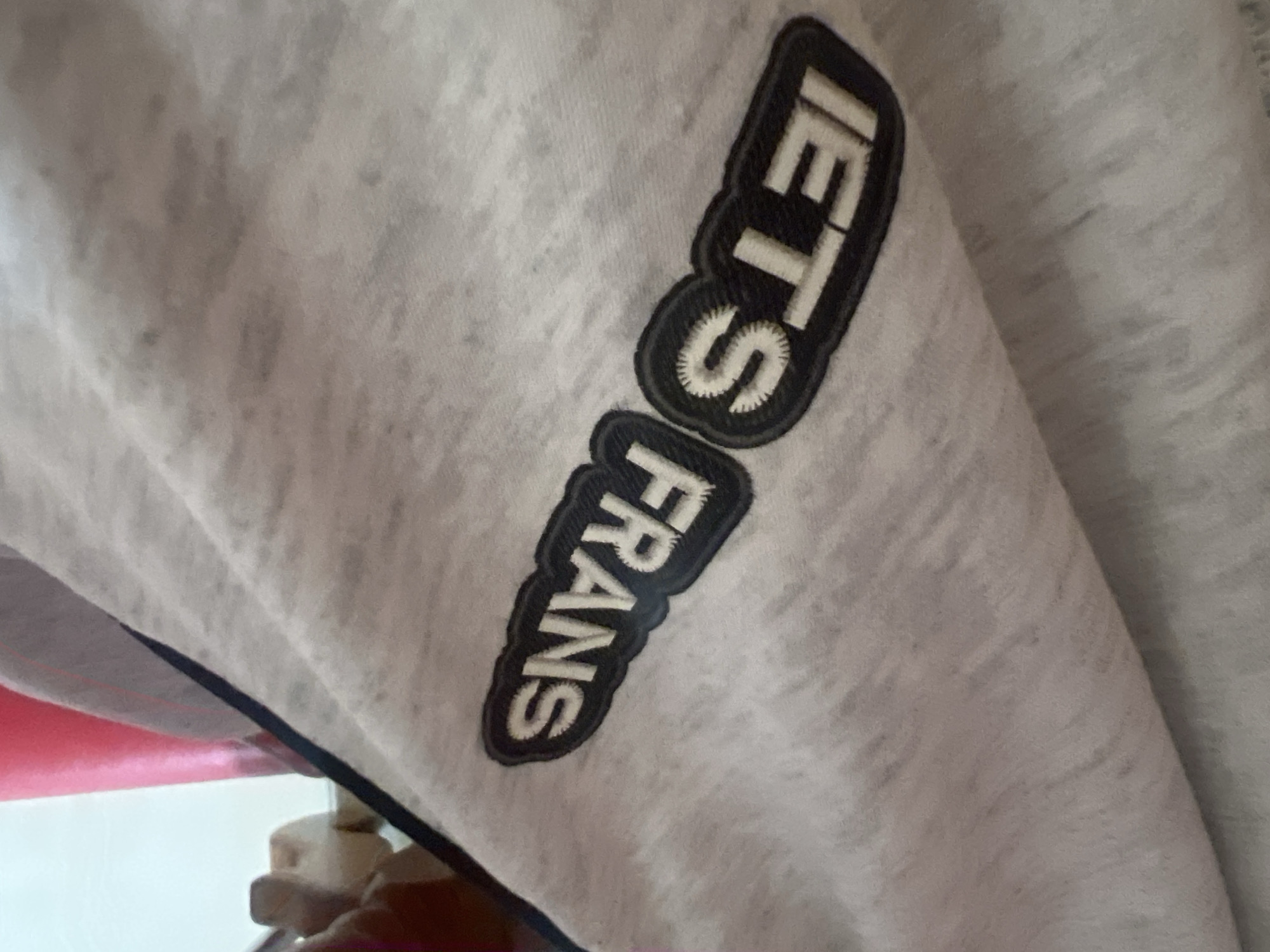 The image displays a close-up view of a light gray fabric, possibly a sweatshirt or t-shirt, with embroidered text. The text is arranged vertically and appears to be a logo or brand name. The top portion of the text reads "IETS", with the letters being white and outlined in black. Below this, the word "FRANS" is visible, also in white letters with a black outline. The embroidery is slightly raised, indicating a textured patch. The fabric itself has a slightly mottled appearance, with subtle variations in shade, suggesting a knit material. In the background, a hint of a red surface and a white object are visible, but they are out of focus and do not provide clear context. The lighting appears to be artificial, casting soft shadows on the fabric. No specific time of day, weather, or location cues are evident, and the overall tone is neutral, focusing solely on the garment's details.