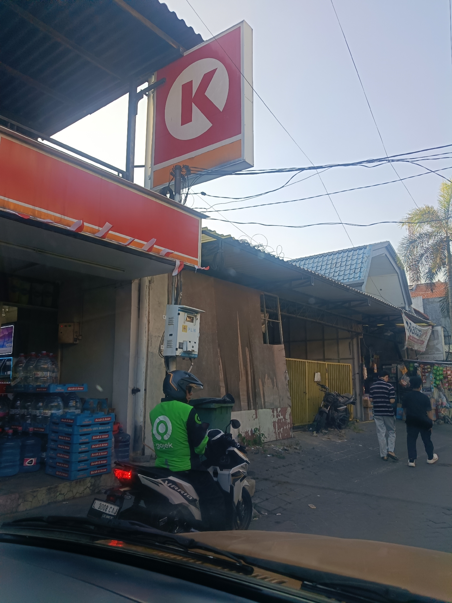 The image captures a street scene in Surabaya, Indonesia, likely near a convenience store. In the foreground, a person wearing a green Gojek jacket and helmet is seated on a scooter, facing away from the camera. The scooter is parked on a paved road. To the left, a convenience store with a red awning is visible, displaying stacks of bottled water. A large red sign with a white "K" is prominently displayed above the store. In the background, several people are walking on the street, and a motorcycle is parked near a yellow gate. Buildings with tiled roofs line the street. The overall environment suggests a bustling commercial area in a tropical climate, as indicated by the palm tree on the right side of the image.