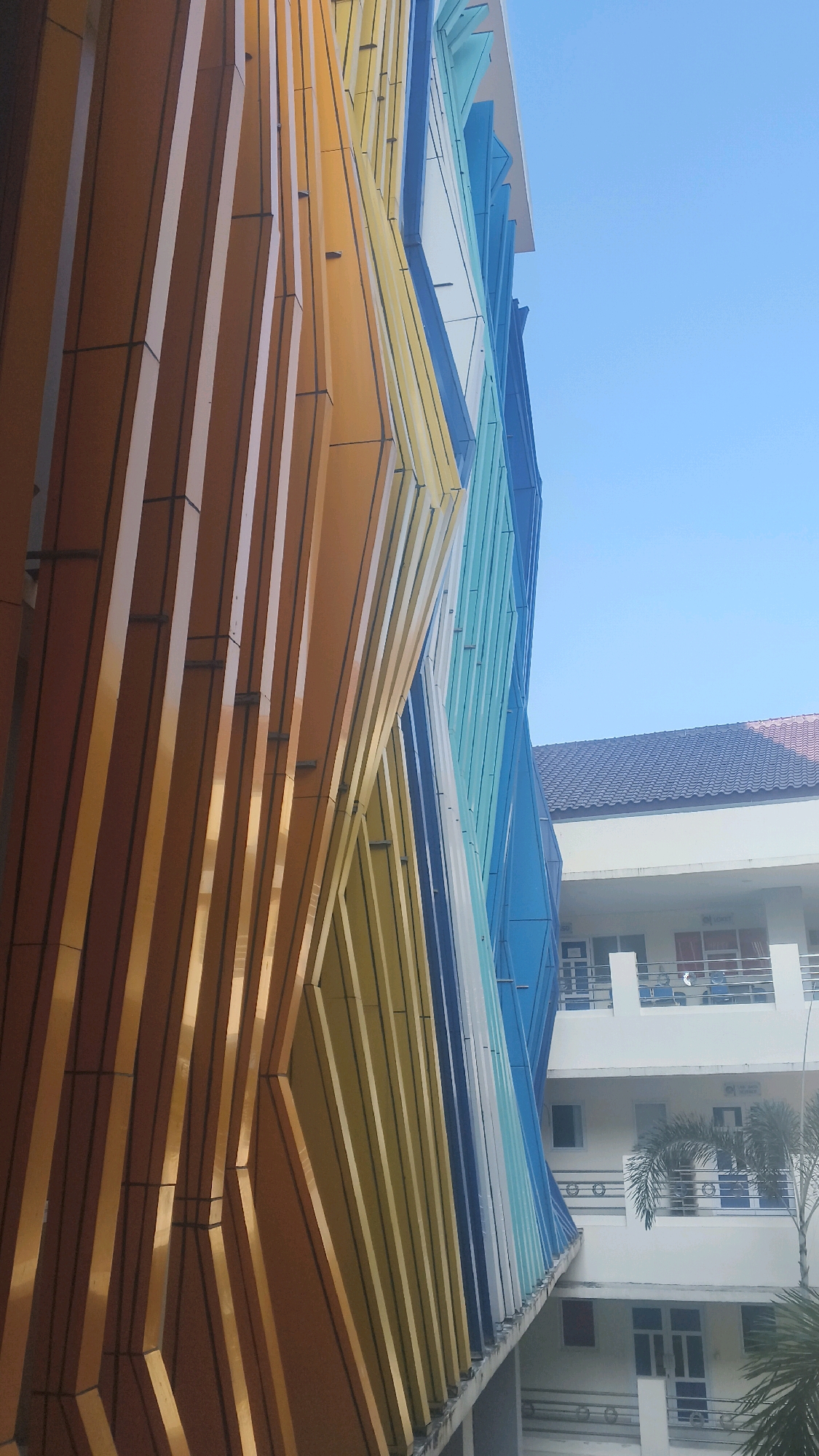The photograph, taken from a low angle, captures the exterior of a modern building in Lohbener, Indonesia. The building's facade is adorned with a striking, colorful design featuring vertical panels that twist and turn, creating a dynamic, undulating effect. The panels transition in color from warm browns and yellows on the left to blues and greens on the right. In the background, another building with a white facade and dark tiled roof is visible. This building has multiple stories with balconies and windows, suggesting it is an institutional or residential structure. A palm tree is partially visible in front of the background building. The sky above is a clear, bright blue. The image appears to have been taken during daylight, with the sun casting reflections on the metallic panels of the facade. There are no people visible in the frame.