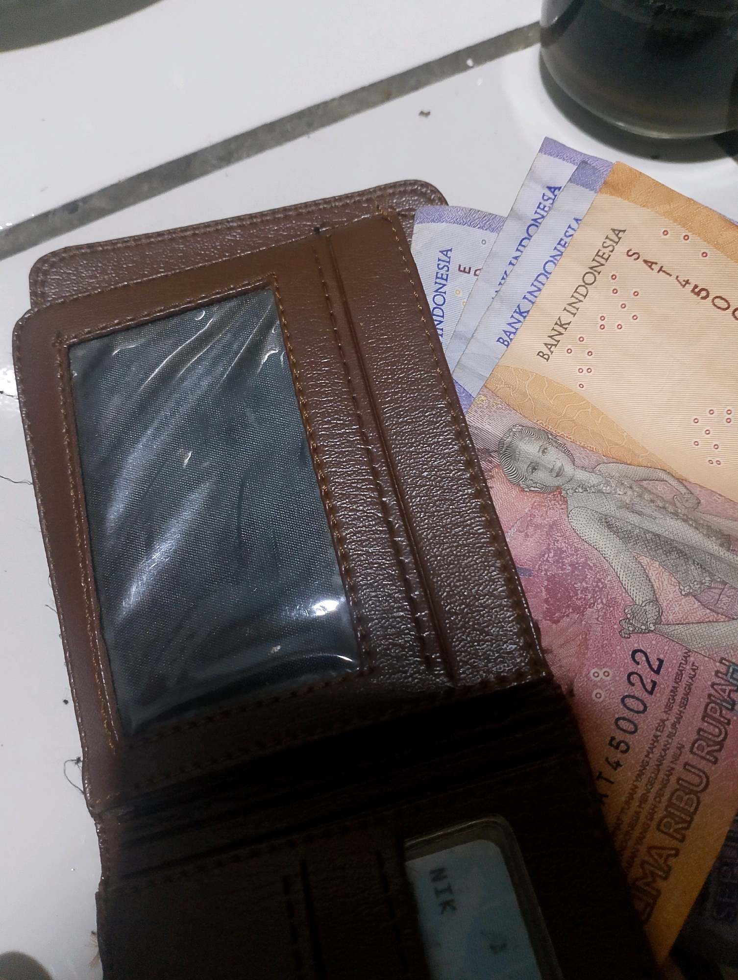 The image shows a brown leather wallet open to reveal several Indonesian Rupiah banknotes. One banknote, a 2,000 Rupiah note, features a portrait of Soesilo Soedarman on the obverse and a depiction of a family on the reverse, along with the text "BANK INDONESIA" and "SATU RIBU RUPIAH" which means "one thousand Rupiah". Another banknote is partially visible with the text "BANK INDONESIA" and "INDONESIA". The wallet also has a transparent window, likely for an identification card. The background appears to be a white surface, possibly a table, with some dark circular objects in the upper right corner. The lighting is somewhat dim, casting shadows. There are no people or animals visible in the image. This scene is not specifically located in the city of Ciomas, Indonesia; it is a generic indoor scene.