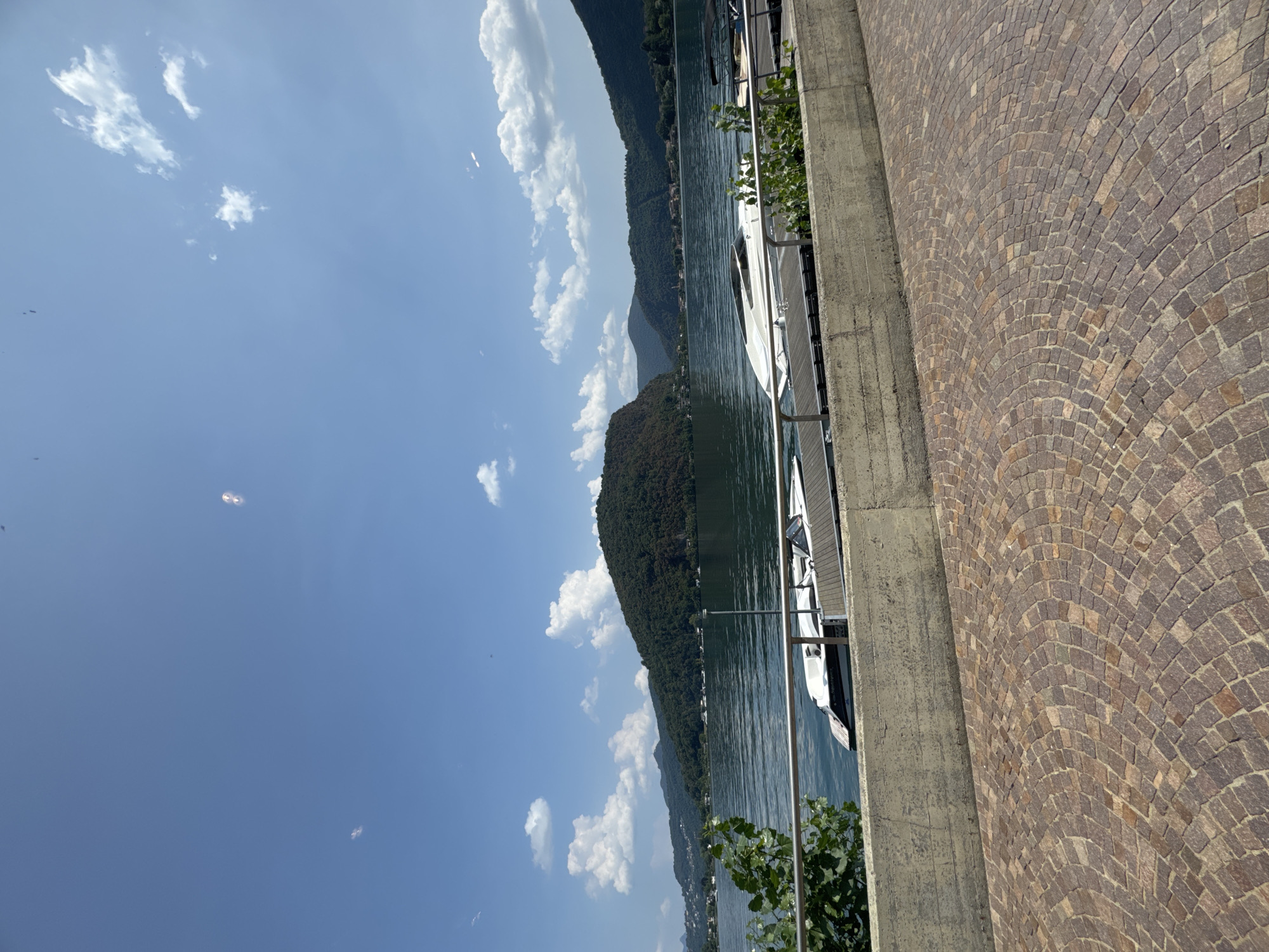 The image shows a view of Lake Lugano from a paved area in the city of Lavena Ponte Tresa, Italy. The lake is calm and reflects the blue sky, which is dotted with white clouds. In the foreground, a stone-paved walkway curves away from the viewer, bordered by a concrete wall. On the other side of the wall, a dock extends into the water, with two white motorboats moored to it. Greenery grows along the edge of the dock and the concrete wall. In the background, lush green hills rise from the opposite shore of the lake, with some buildings visible nestled amongst the trees. The overall scene depicts a sunny day at a scenic lakeside location.