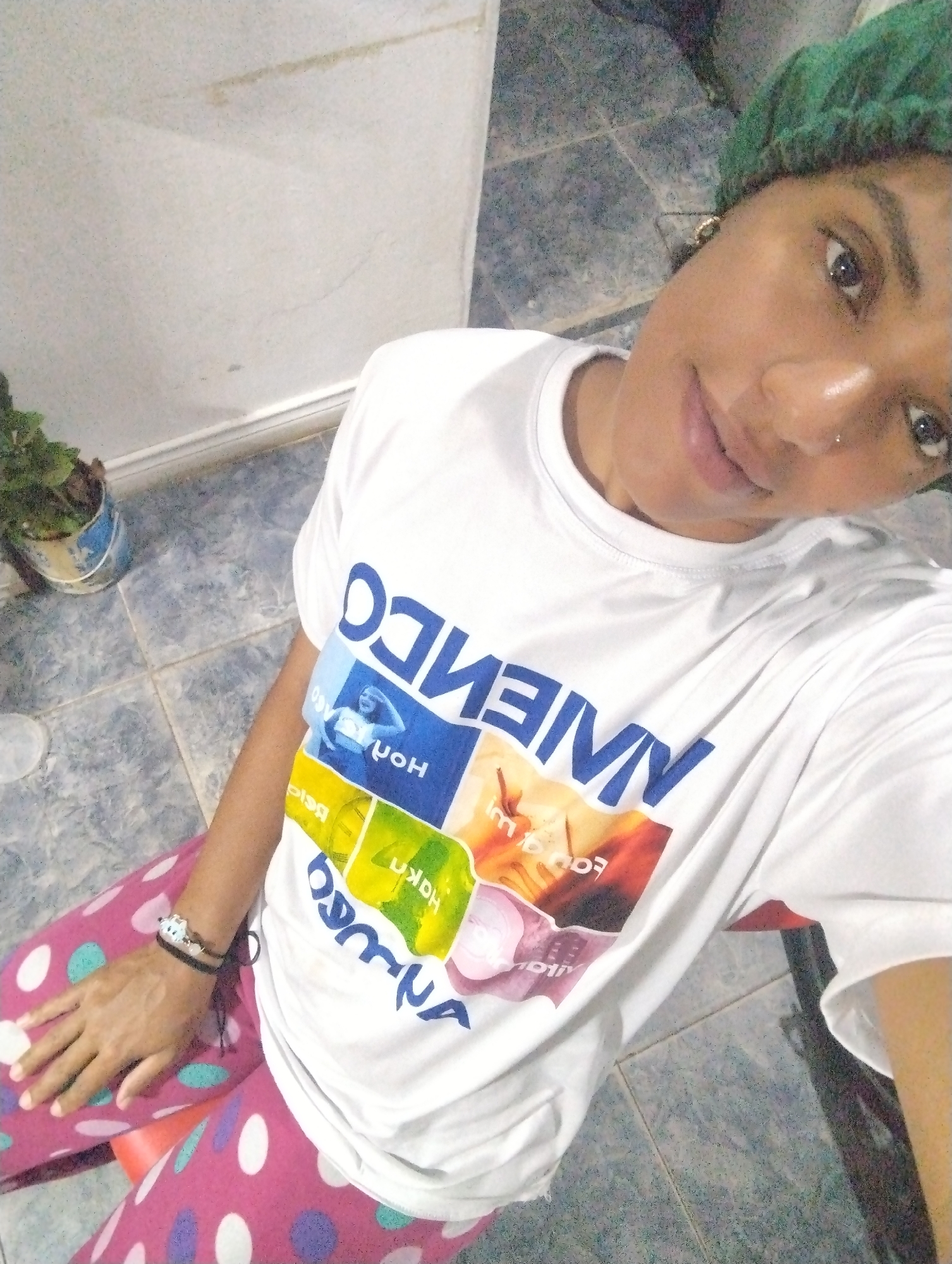 A medium, high-angle shot captures a woman wearing a green hair covering, a white t-shirt with colorful graphics, and polka-dotted pants, likely indoors on a tiled floor. A potted plant is visible in the background. The scene does not provide information to verify the location in the city of El Valle del Espíritu Santo, Venezuela.