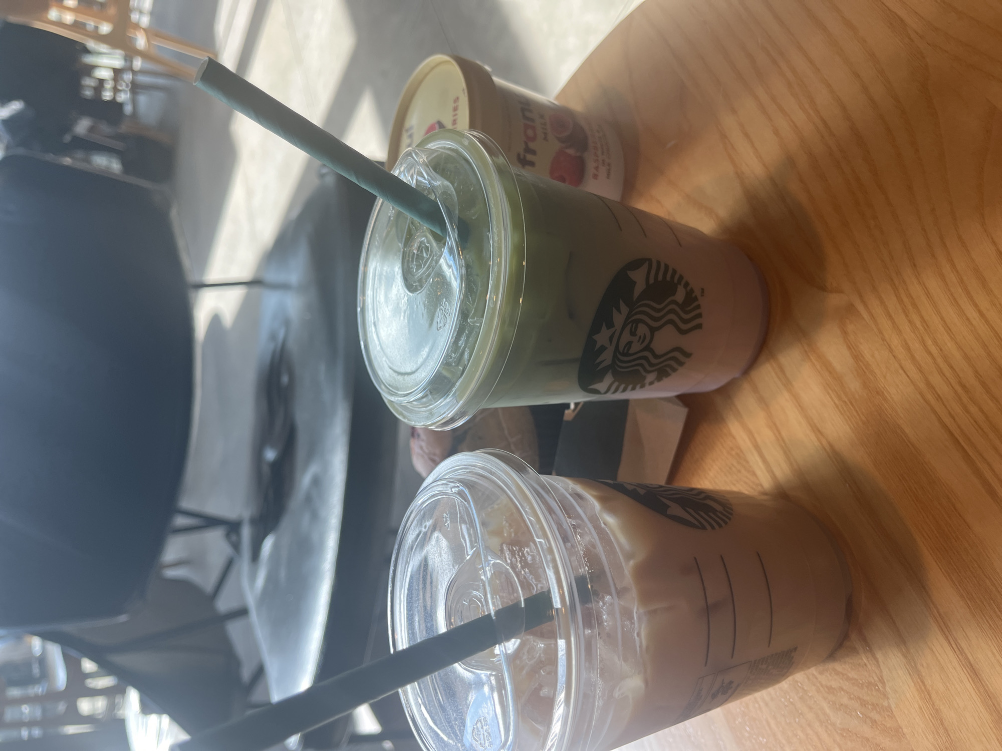 The image captures a casual tabletop scene, likely within a Starbucks cafe in Plovdiv, Bulgaria, during daytime. The prominent subjects are two clear plastic iced beverage cups, both featuring the recognizable Starbucks mermaid logo.
One cup, positioned further back, contains a layered green drink, consistent with an iced matcha latte, and has a green paper straw. Next to it, closer to the viewer, is another cup holding a light, creamy brown iced beverage, likely an iced coffee or latte, with a dark grey paper straw. Both cups exhibit condensation, indicating their cold temperature.
Partially visible behind the green drink is a small, round, beige container with a red label. Legible text on the label includes "fran" (possibly part of a brand name), "MILK," and "RASPBERRIES," suggesting it is a yogurt or dessert product.
These items are set on a light-colored wooden table, characterized by visible grain patterns. The scene is illuminated by bright natural sunlight, which casts distinct shadows from the cups and straws onto the table, implying a sunny day. The background is softly blurred and dark, showing indistinct shapes that could be other cafe furniture or interior elements. No people are visible in the frame. The overall impression is one of a relaxed break, enjoying drinks and a snack in a brightly lit cafe setting.