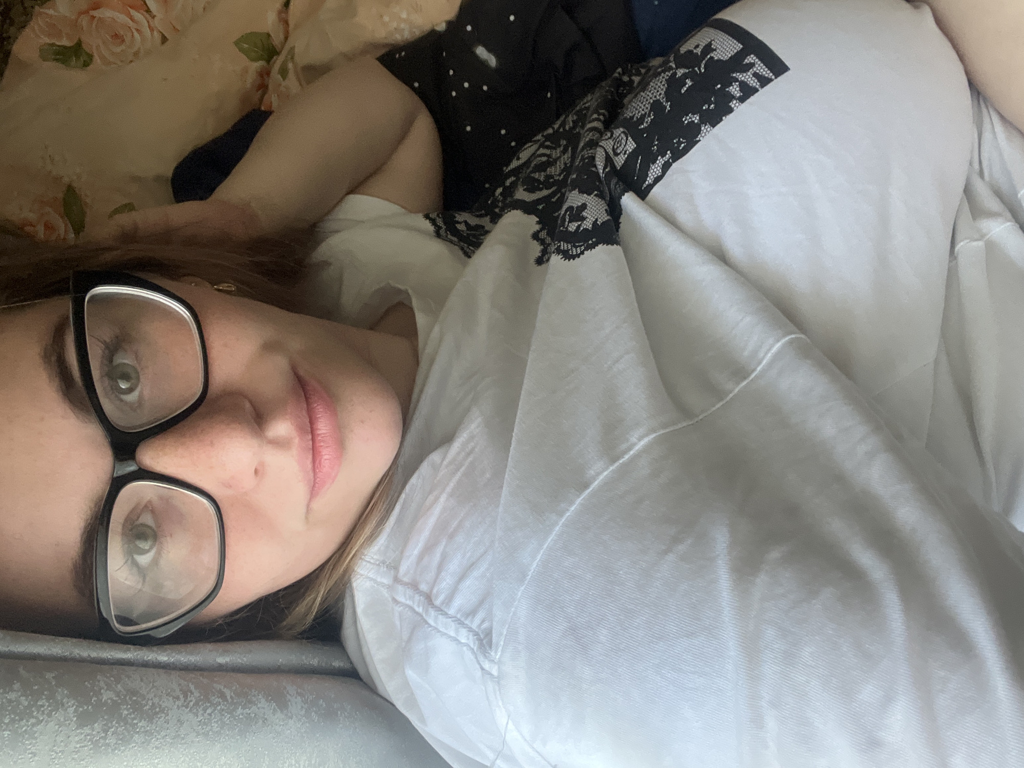 A woman with glasses is lying on a bed. The bed has a floral patterned sheet and a light grey pillow. She is wearing a white t-shirt with black lace on the shoulder. Another piece of clothing, black with white polka dots, is visible behind her. The location is unknown, and there is no evidence to suggest it is in the city of Chaikovsky, Russia.
