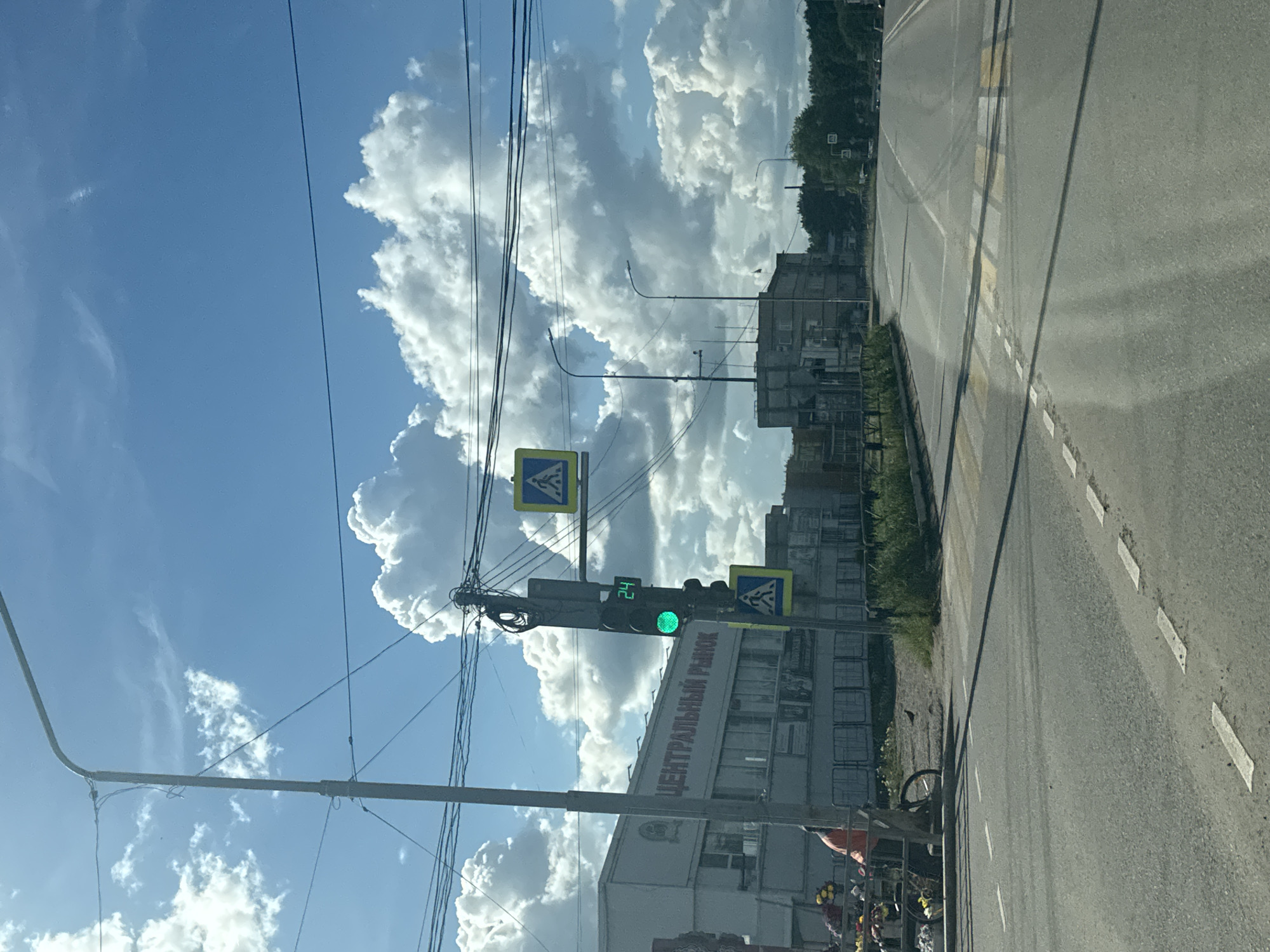 The image depicts a street scene in Chaikovsky, Russia. The road is on the right side of the image. The sky is prominently visible, filled with fluffy white cumulus clouds against a blue backdrop. Power lines crisscross the sky above a crosswalk with a green light indicating it is safe to cross. The building to the left bears the inscription "ЦЕНТРАЛЬНЫЙ РЫНОК" which translates to "Central Market" in English. In front of the building, on the right side of the road, there is what appears to be a floral vendor near the side of the road. A bicycle is resting near the side of the road. In the distance, there are buildings.