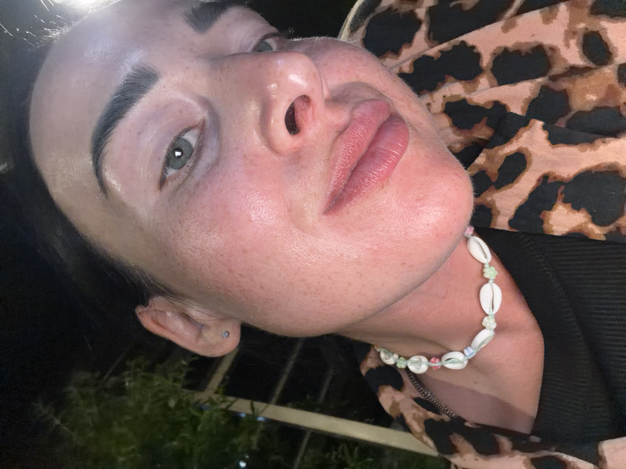 A close-up, low-angle selfie taken at night shows a person's face and upper body against a dark background with some foliage and a vertical element that could be a building in Sochi, Russia. The person, who appears to be female with dark hair and light skin with freckles, is wearing a black ribbed tank top and a leopard-print garment. A white shell and bead necklace is visible around her neck. Her left eye, light green or grey, is visible along with her brow and part of her forehead. Her nose and lips are also prominently featured. The lighting suggests artificial light sources, possibly from a nearby establishment or streetlights, creating some highlights on her skin.