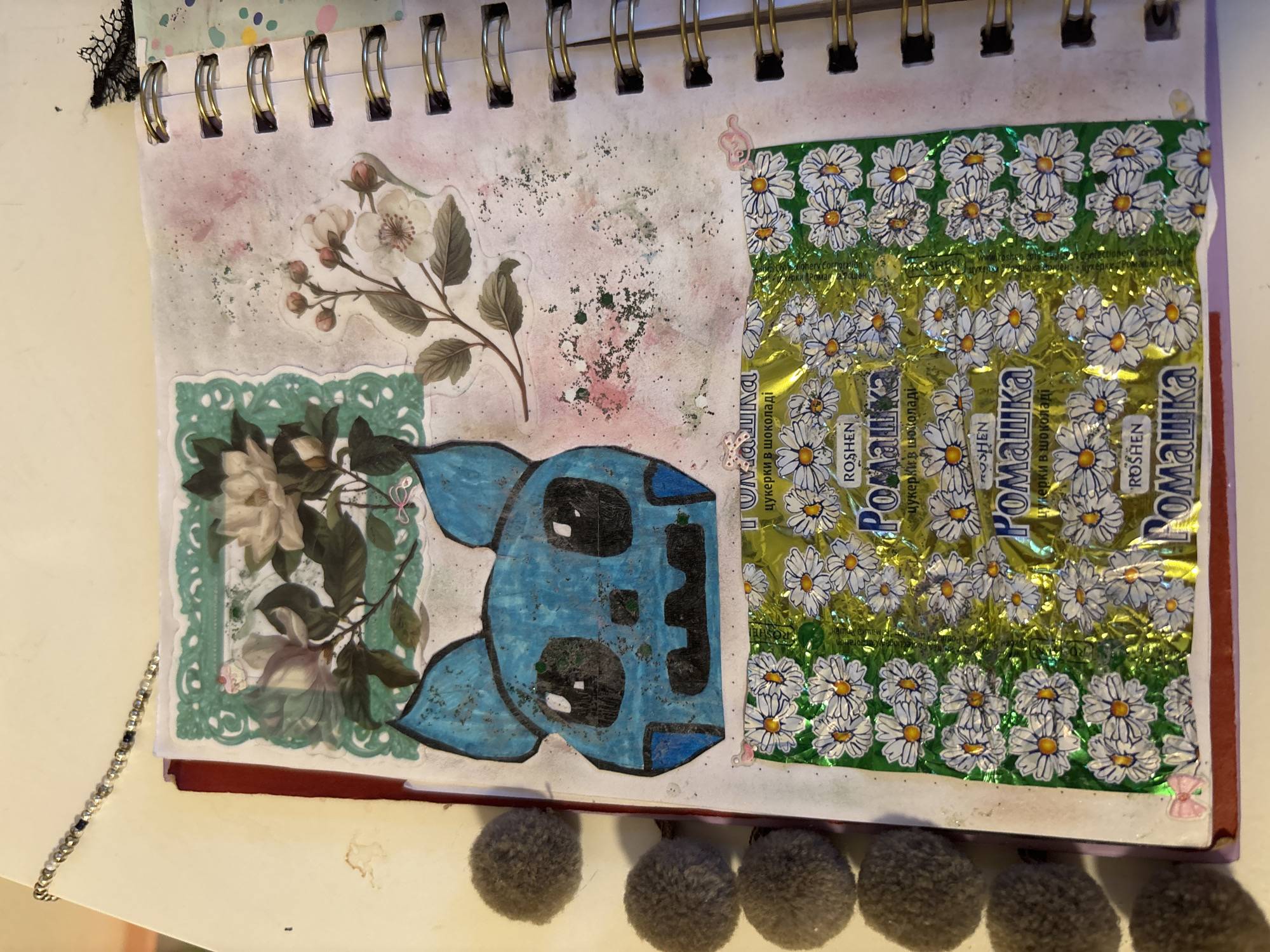 The image shows a close-up view of a scrapbook or journal page. The page is decorated with various elements, including stickers, drawings, and a candy wrapper.
On the upper portion of the page, there are two sticker embellishments: one depicts white flowers and green leaves within a decorative green border, and the other shows a branch with pink-tinged white flowers and buds. In the center of the page, a blue, cartoon-like creature with large black eyes, ears, and a grimacing mouth is drawn.
Below the drawing, a foil candy wrapper is attached to the page. The wrapper is predominantly green and gold and features a repeating pattern of white daisies with yellow centers. Text in Cyrillic script is visible on the wrapper.
To the left of the journal page, a string of small, round, gray pom-poms is attached. The journal itself appears to be a spiral-bound notebook with a dark red cover visible at the edge. The background surrounding the journal page is a neutral, light-colored surface.