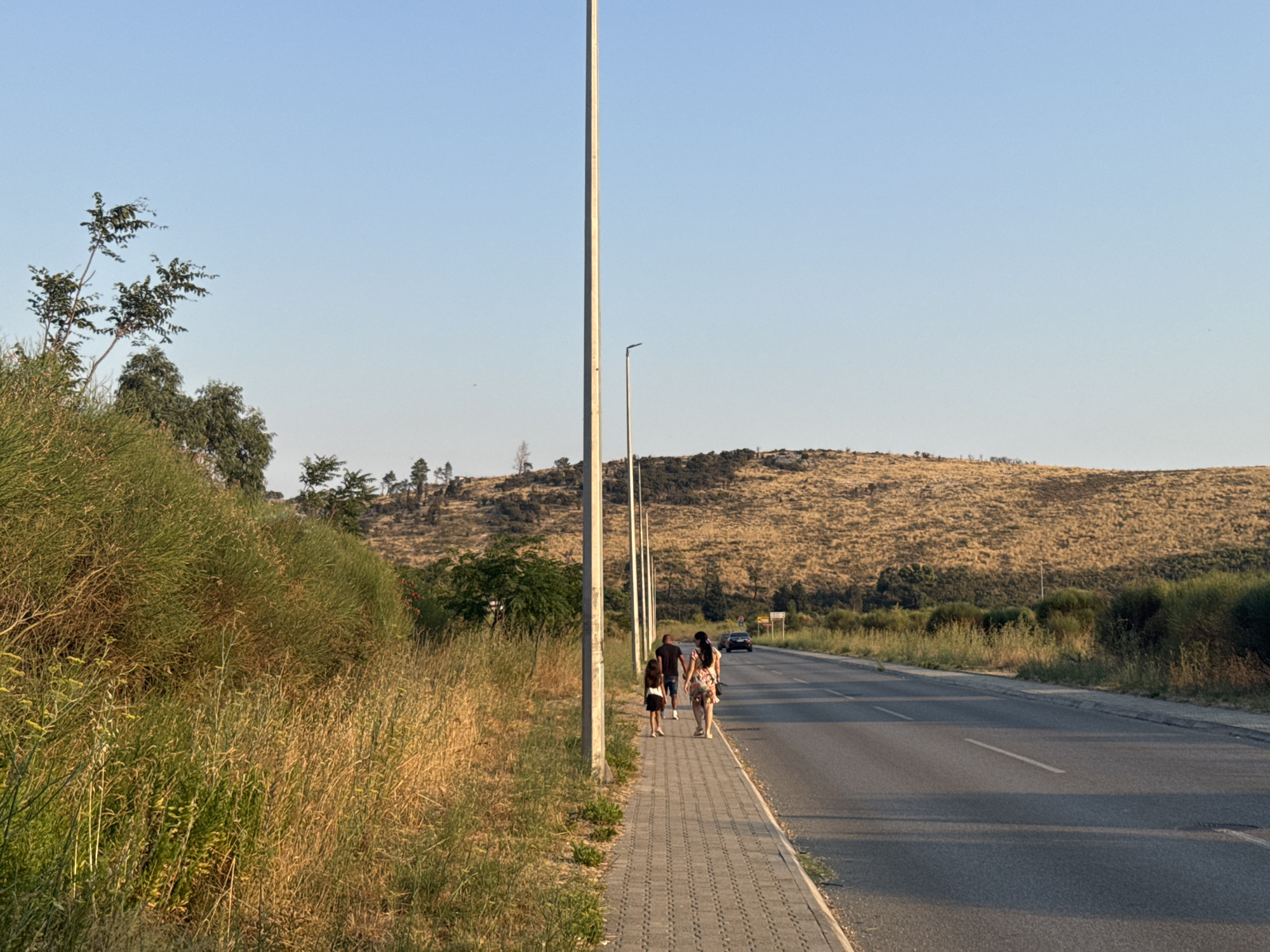 The image depicts a wide paved road with a pedestrian walkway on the left. The walkway is bordered by dense, dry grass and bushes. Several tall streetlights are spaced along the walkway. In the background, a barren, golden-brown hillside rises, dotted with sparse trees and shrubs.
Four individuals are walking away from the camera on the pedestrian walkway. The first two are children, one wearing a dark shirt and skirt, and the second, slightly ahead, is wearing a patterned dress. Behind them, two adults are walking; one in a dark t-shirt and shorts, and the other in a light-colored, floral patterned dress. The group appears to be walking towards the right side of the image, away from the viewer.
A dark-colored car is visible further down the road, moving in the same direction as the pedestrians. The sky is clear and a light blue. The overall scene suggests a rural or suburban setting on a clear day, possibly in the late afternoon due to the warm lighting on the hillside.
This scene is located on the main road entering Sutomore, Montenegro from the direction of Bar. The landscape with the characteristic dry, rocky hills is typical for this coastal region of Montenegro.
