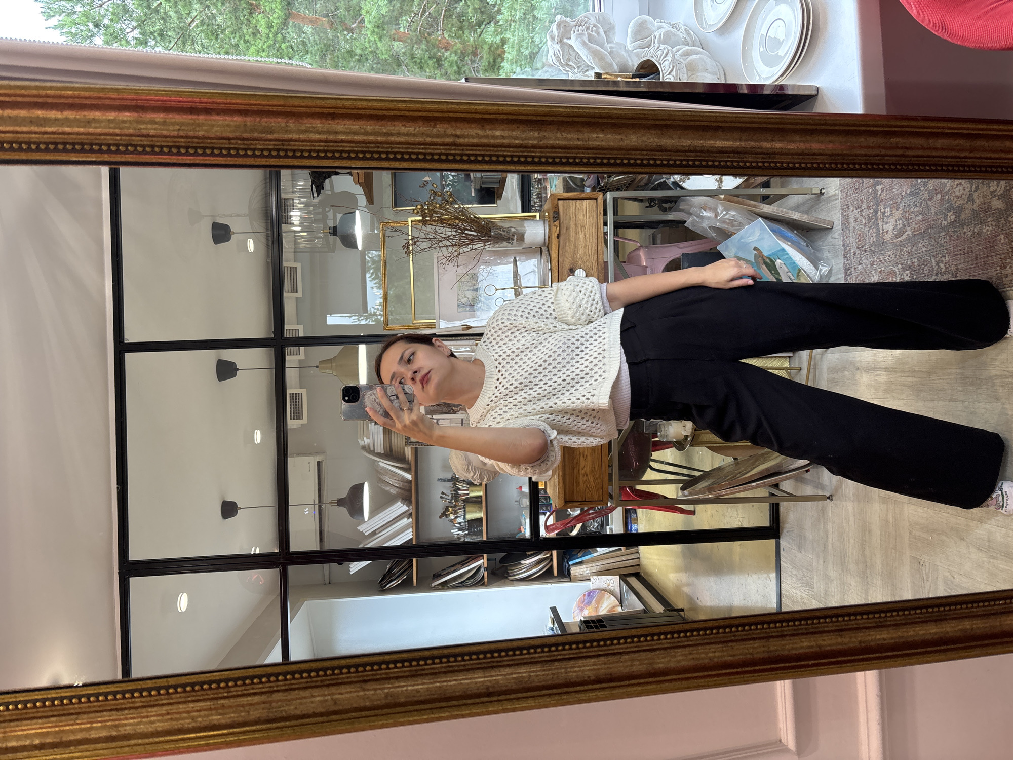 A young woman stands in a room with a large ornate mirror, taking a selfie. She is wearing a white, open-knit sweater and black wide-leg pants. The mirror reflects the room behind her, which appears to be an art studio or shop. Shelves are filled with canvases, art supplies, and decorative items. A large window on the right side of the frame looks out onto trees. The woman is focused on her phone, and her expression is neutral. There are no other people visible in the frame. The setting appears to be indoors, in a well-lit space with a mix of modern and antique elements.