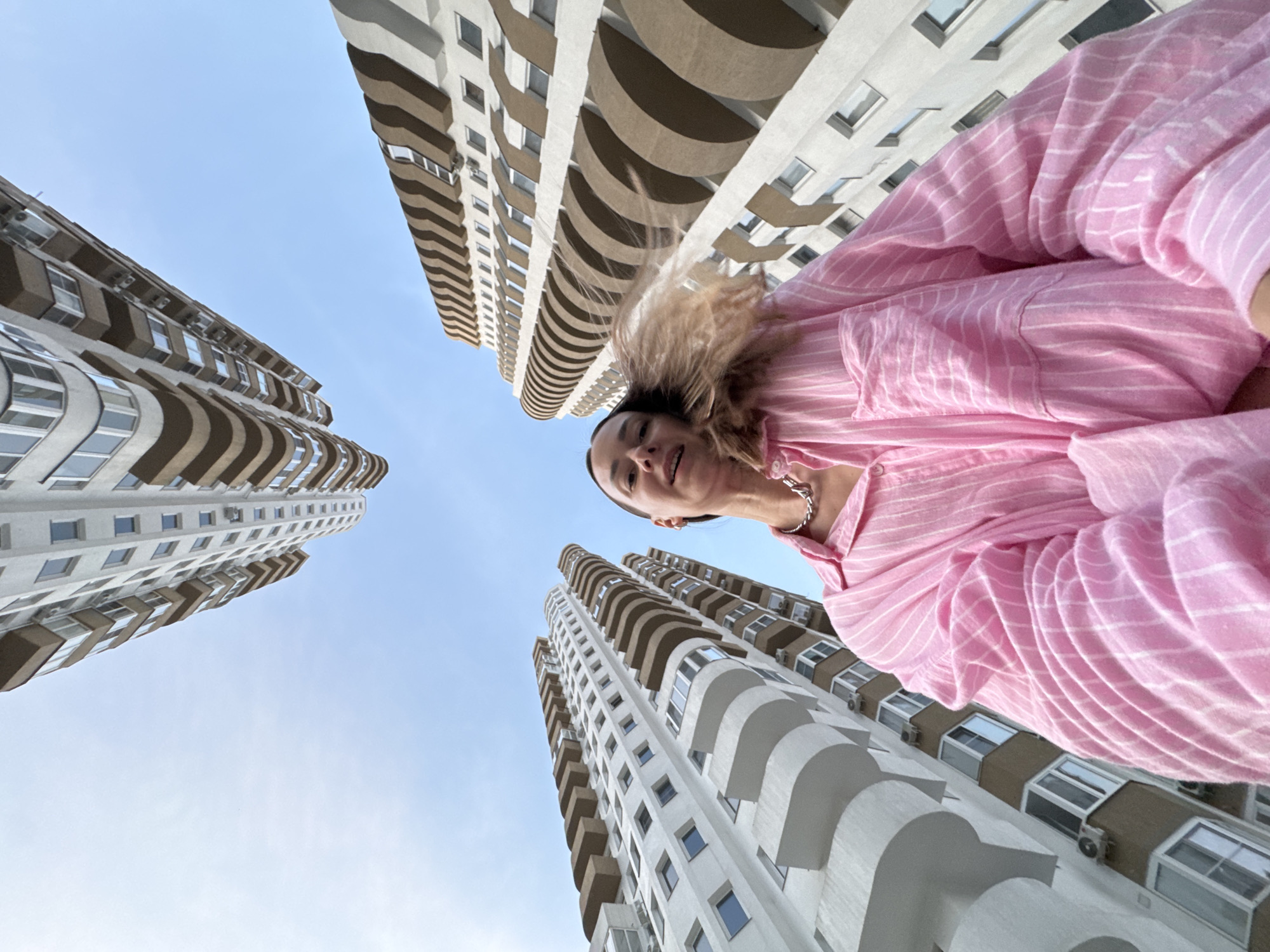 A low-angle, wide-shot photograph captures a young woman standing in an urban environment, with residential buildings dominating the background. The woman, positioned in the foreground, is looking upwards and slightly towards the camera. She is wearing a pink and white striped oversized shirt and a silver necklace. Her hair is light brown and appears to be blowing in the wind. Multiple tall, modern residential buildings with balconies are visible, appearing to surround her. The sky is clear and blue, suggesting it is daytime. The architectural style of the buildings, with their curved balconies and white facades, is characteristic of recent construction. There is no clear indication of the specific city in the image itself, however, the provided context states the location is Sochi, Russia.
