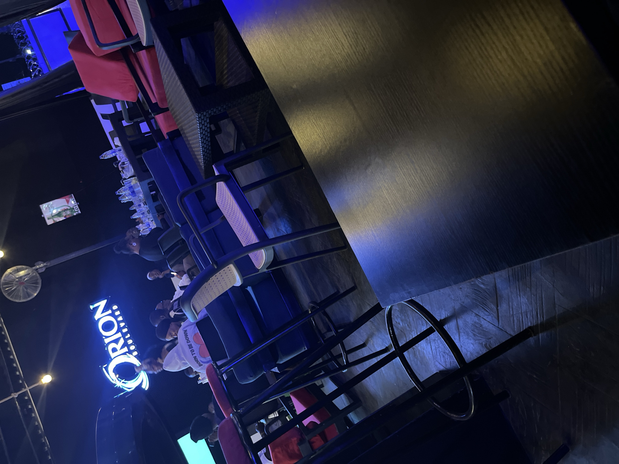 The image depicts the interior of a dimly lit establishment, possibly a lounge or club, with blue and pink neon lighting. A prominent "ORION BOULEVARD" sign is visible, illuminated in blue. Several individuals are seated at tables and chairs, some engaged in conversation. The furniture includes blue sofas, red cushioned chairs, and dark-colored tables with woven textures. Bar stools with pink seats are also present. In the background, a large screen displays an advertisement. The floor appears to be tiled with a wood-grain pattern. The overall ambiance suggests a social gathering or entertainment venue. The establishment is located in Abuja, Nigeria.