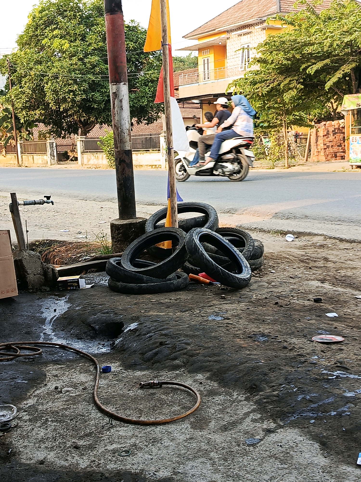 In Megang Sakti I, Indonesia, a scene unfolds on a roadside, capturing a moment near a motorcycle repair or service area. A weathered red and white pole stands near the foreground, adorned with colorful flags, suggesting a festive or market atmosphere. To the left of the pole, a faucet attached to a pipe is visible, hinting at water access for the area's operations.
Several worn motorcycle tires are piled haphazardly on the ground, some stacked and others scattered, indicating their used condition. The ground is uneven and appears to be a mix of dirt, mud, and possibly oil or grease, consistent with a workspace. A rusty hosepipe lies coiled on the ground, extending towards the lower left corner of the frame, with a nozzle attached at its end.
In the background, a white motorcycle is in motion, carrying two individuals. The person in the rear, a woman, is wearing a light-colored hijab and a patterned top with blue jeans. The person in the front, a man, wears a dark t-shirt and a white cap. They are moving away from the camera, towards a street that borders the property.
Beyond the street, a yellow, two-story building with white window frames stands prominently, its architecture suggesting a residential or commercial structure. Adjacent to this building, a vibrant green tree with dense foliage provides a natural backdrop. Further to the right, a wall constructed of reddish-brown bricks is visible, adding texture to the environment. The overall lighting suggests it is daytime, with sunlight illuminating the scene.