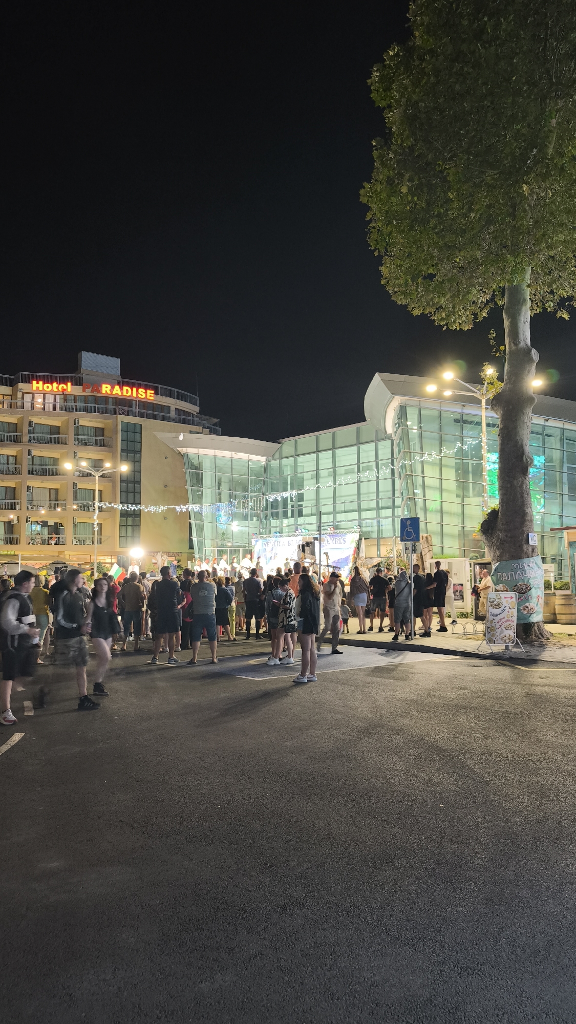 The image captures an outdoor gathering at night in the city of Pomorie, Bulgaria. A large crowd of people stands on an asphalt surface in front of a modern, glass-fronted building that appears to be a commercial establishment or entertainment venue. The building is illuminated, with string lights decorating its facade. To the left, a multi-story hotel, "Hotel PARADISE," is visible, also lit up. A large tree with green foliage stands to the right of the frame, its trunk illuminated by streetlights. Several signs are present, including one on the tree with Cyrillic text and an image of food, and another to the right of the building with a blue accessibility symbol. The gathering seems to be taking place on a street or plaza, with some people holding flags, possibly indicating a protest or a celebration. The overall atmosphere is lively, with the crowd suggesting an event is underway.