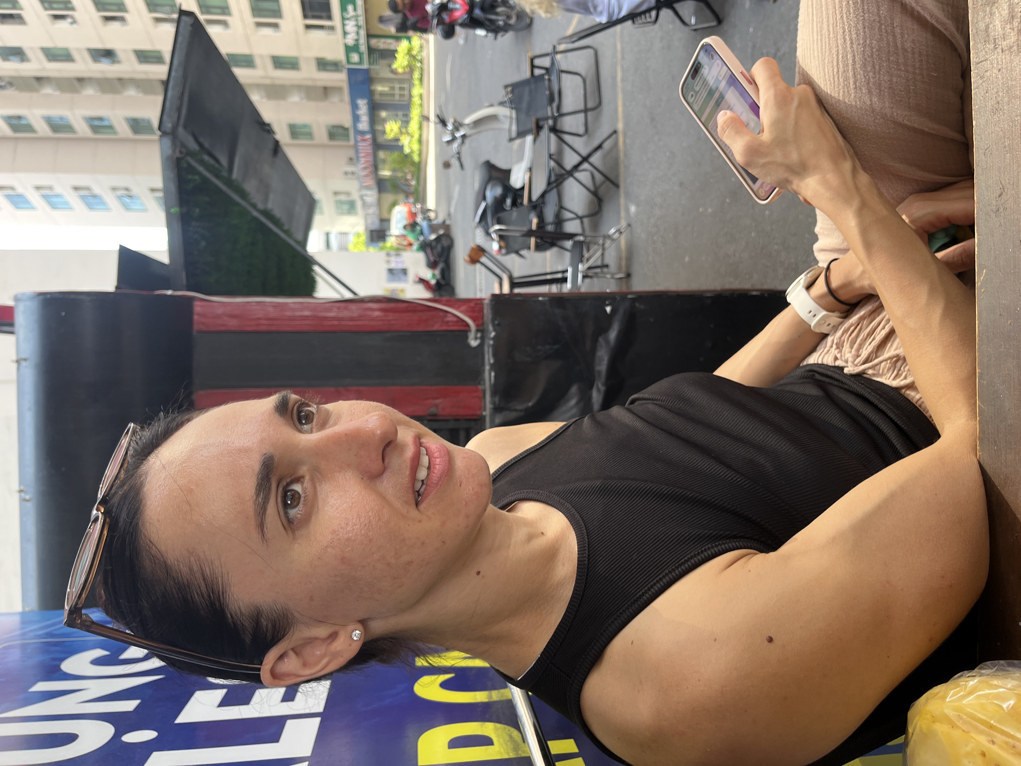 A woman is seated in an outdoor setting, holding a smartphone and looking slightly upward. She is wearing a black tank top and a white smartwatch on her left wrist. A pair of eyeglasses is positioned on her head. In the background, there is a street scene with motorbikes parked and chairs and tables arranged as if for outdoor seating. There's an awning-like structure and signage visible. The image appears to be taken in Bac Nha Trang Ward, Vietnam.