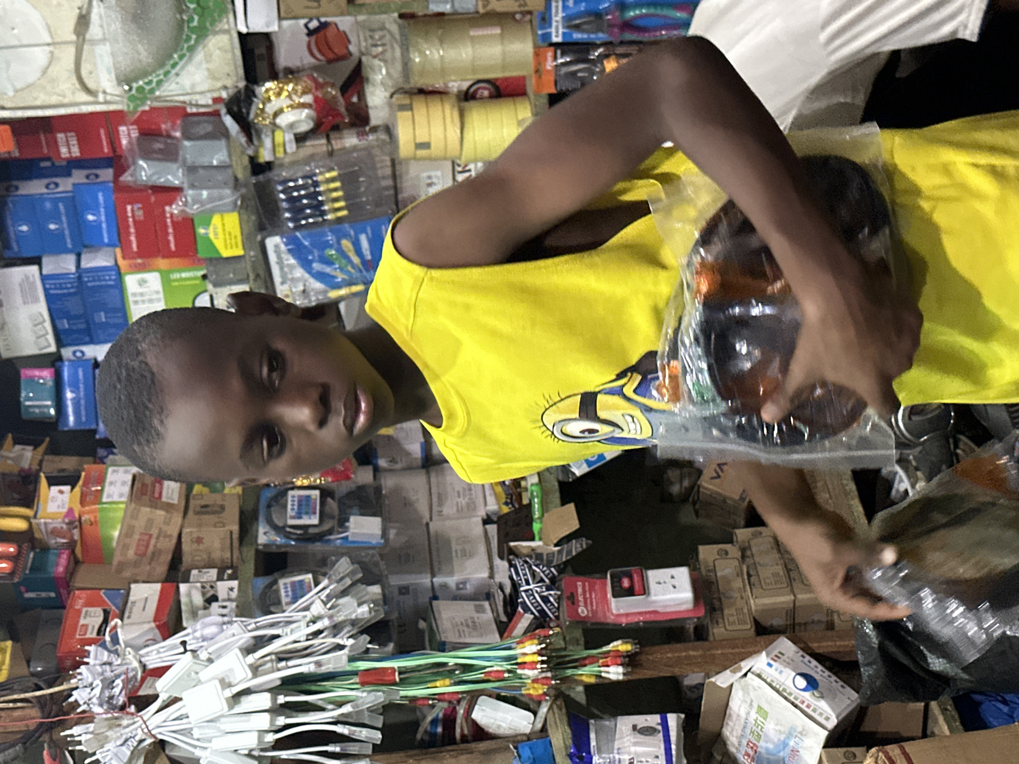 A young boy in a yellow shirt with a Minion graphic stands in a cluttered shop in Barden Yana, Nigeria. He holds a packaged dark brown circular object. The shop is filled with various goods including cables with red, yellow, and white connectors, boxes of electronics, rolls of tape, and tools. The lighting is dim, and the overall impression is of a busy marketplace stall.